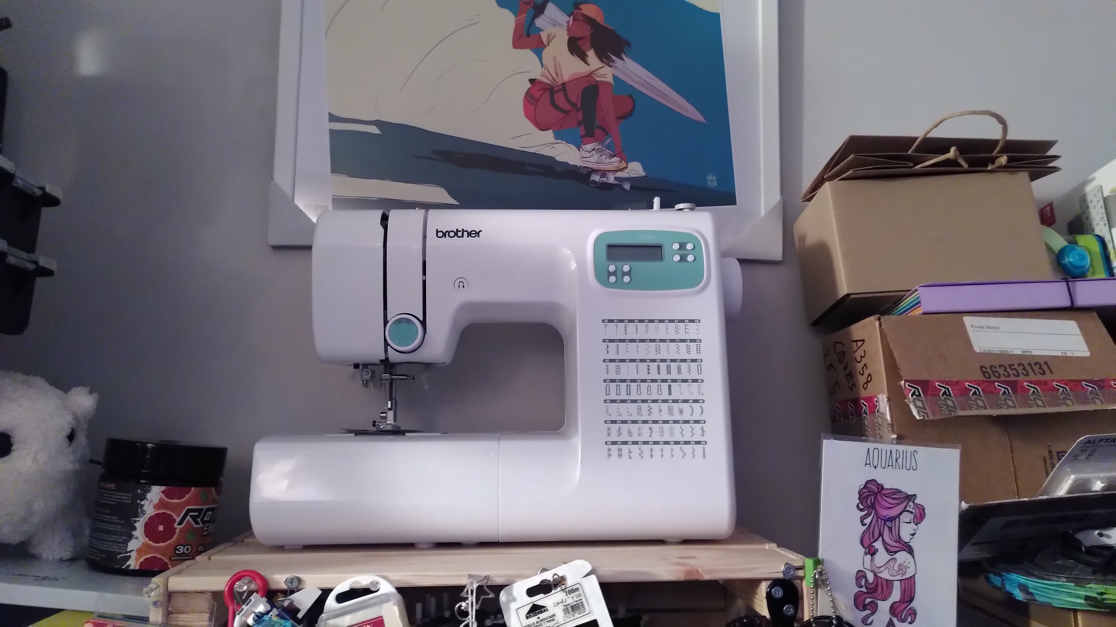 A sewing machine, a brother CS70s. It's white with a small LCD screen with a couple of blue green highlight. It's surrounded by lot of stuff like a print of a skater woman with a big sword by Sara Alfageeh, a print of a girl with pink hair and an aquarius tatoo by heatherjoanneart, a baba plushie...