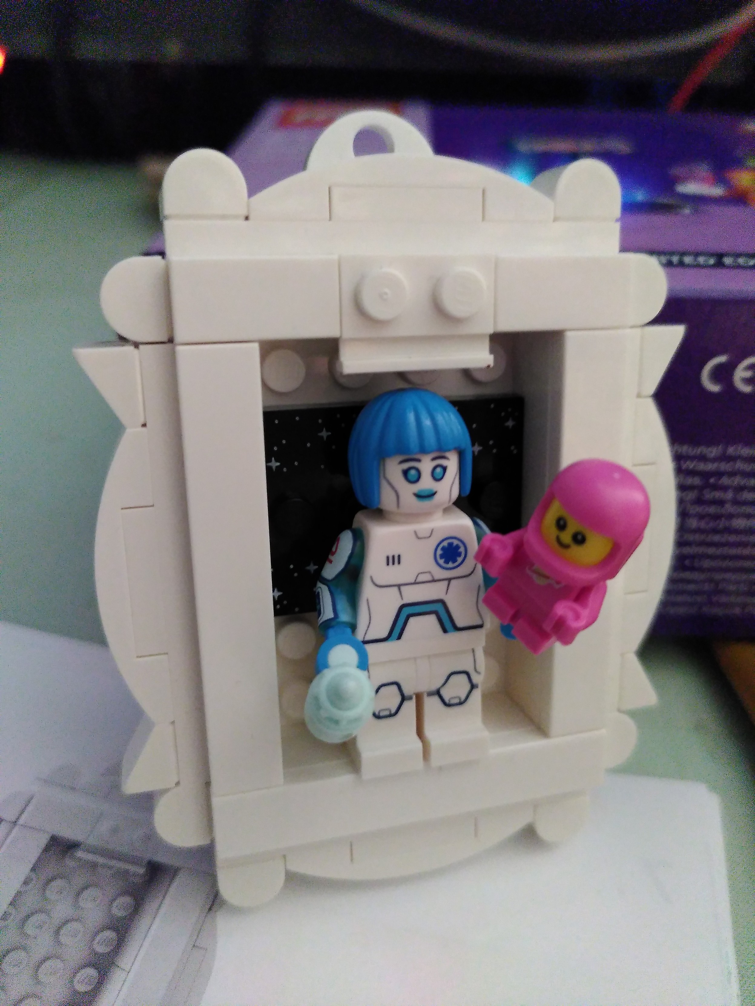 A lego minifigurine representing a robot nurse with blue hair holding a baby in a pink space suit. They are attached inside a frame made of lego