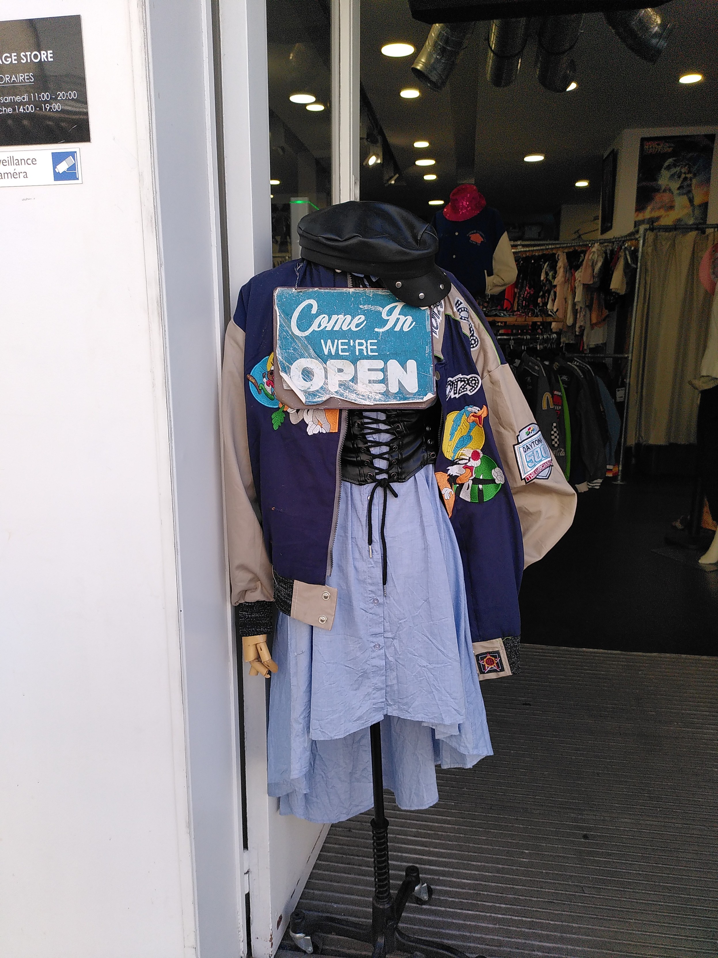 A mannequin in front of a 2nd hand clothes shop. It is wearing a sky blue long shirt; a light black leather corset; a varsity jacket with navy front, sand colored sleeves and patches of speedy gonzales, bugs bunny, the roadrunner and grosminet (the cat who tries to eat the yellow birdie); a leather cap. 