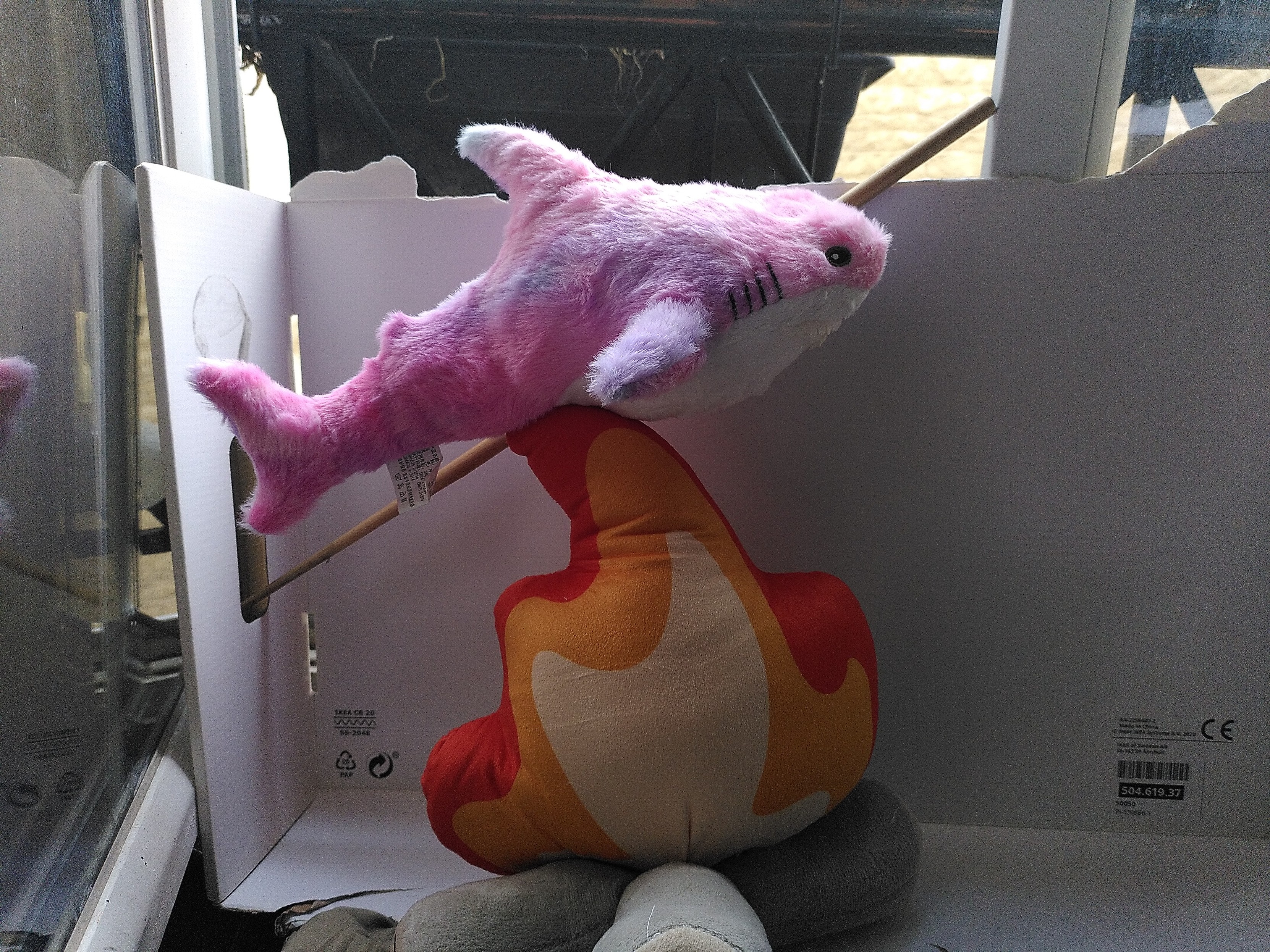 A pink and purple chinese copy of a blahaj attached to a stick above a firewood plushie