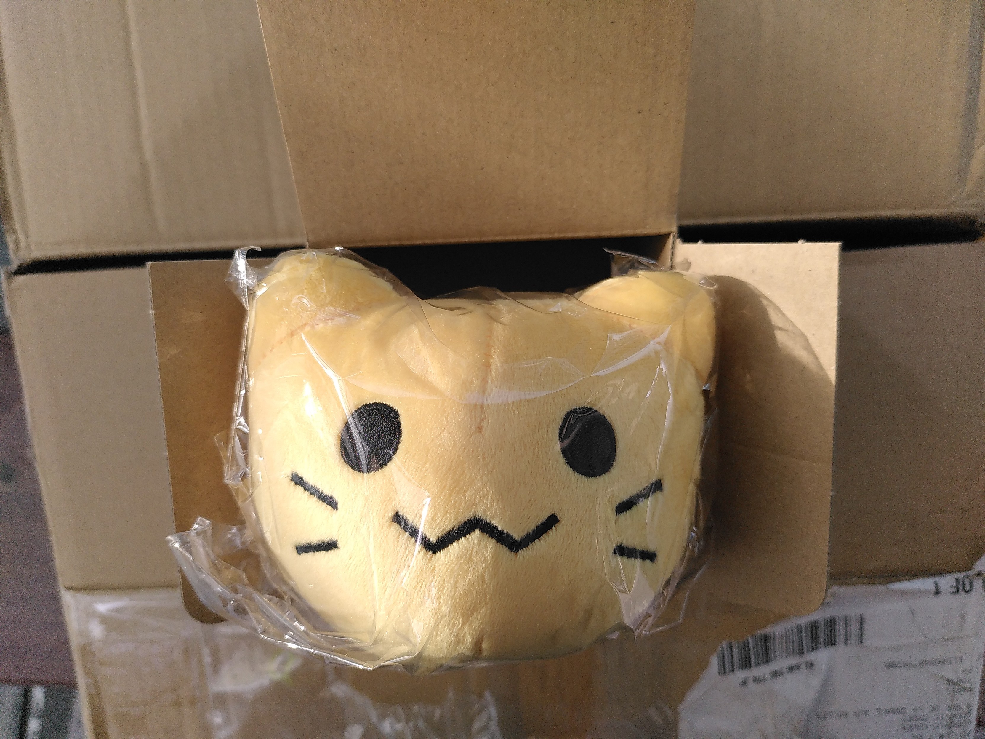 A plushie looking like a yellow blob cat in a plastic wrapper on some cardboard