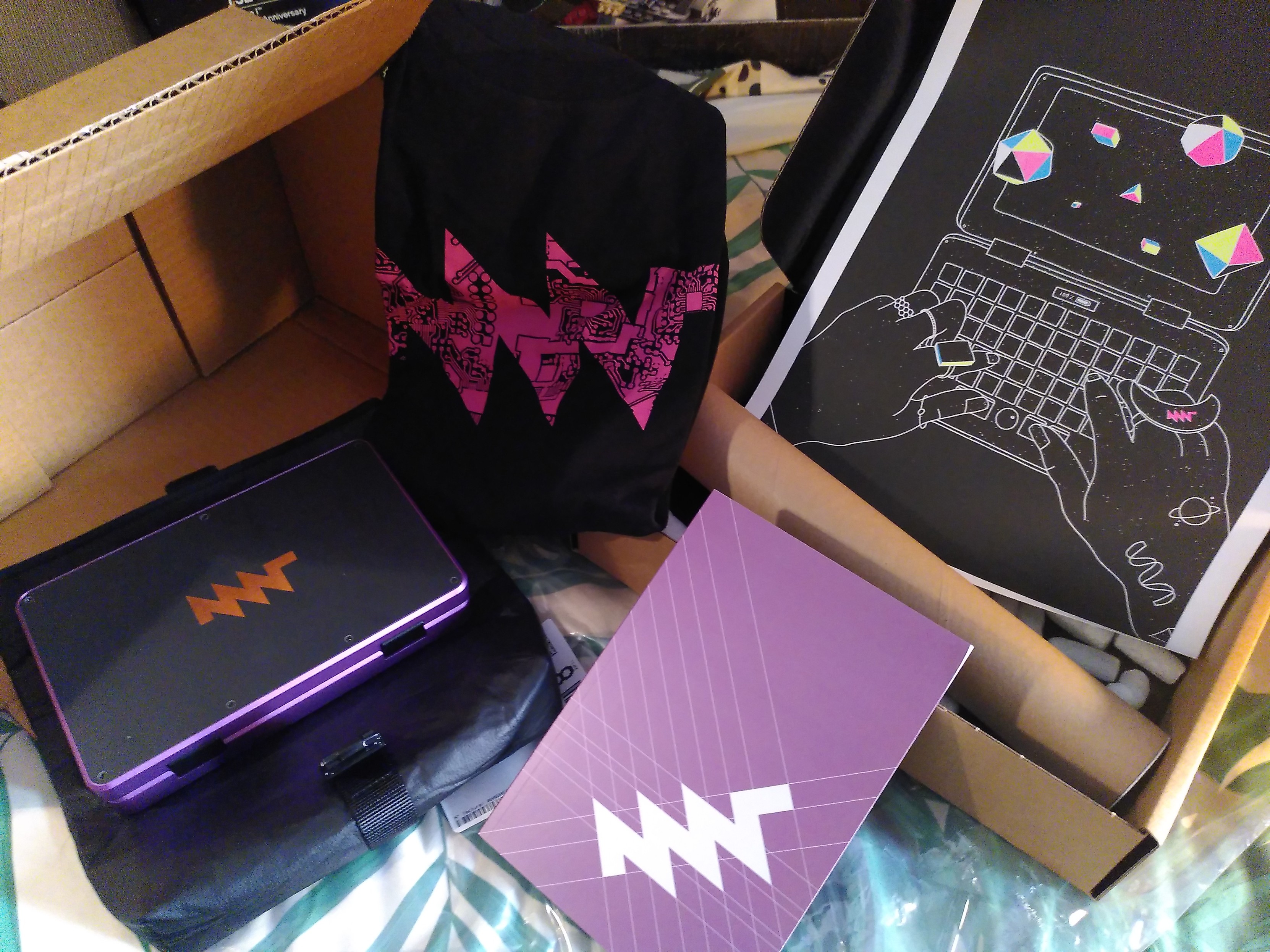 A purple MNT reform pocket - Hyper edition, composed of small computer in pink-purple anodyzed aluminium and black PCB with a large MNT logo made of exposed copper that's small enough to fit in my every day bag, maybe in the front pocket of an eastpack bag; a bag for the computer made of black piñatex, a cloth made of pineapple leaf with a texture similar to leather; a copy of the operator handbook with a purple cover; a A3 print with on one side artwork depicting lineart of the computer and a pair of hand adorned with esoteric jewelry and geometric volume coming out of the screen with each face made of yellow, magenta, cyan or black; a black teesh with the MNT logo in pink PCB pattern that's not part of the hyper edition 