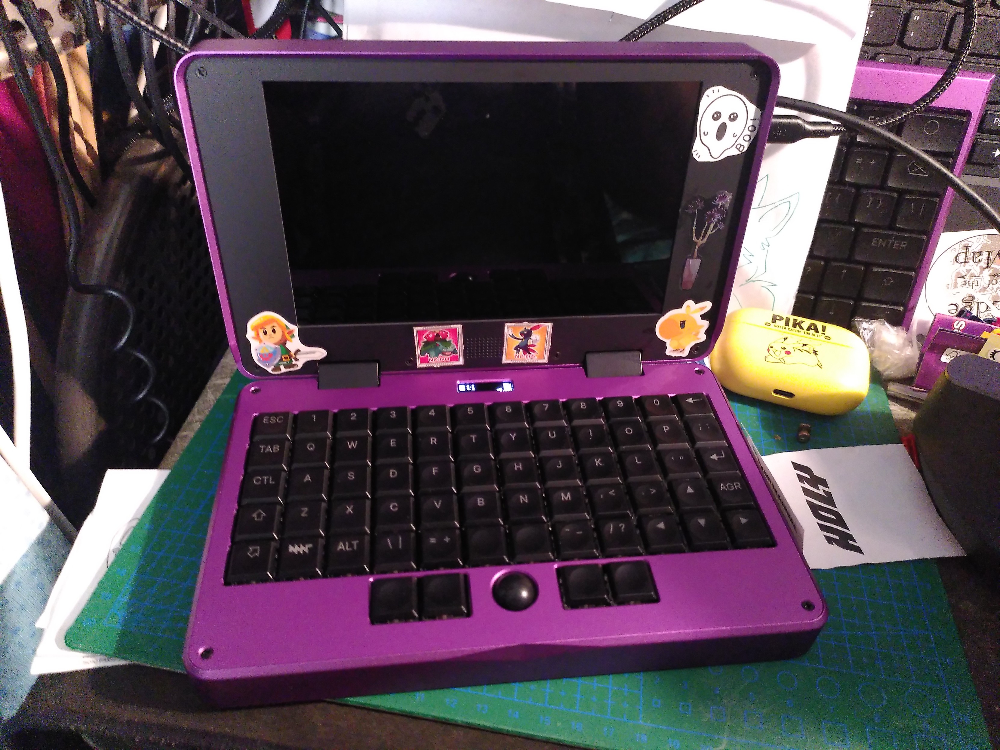 An open MNT reform pocket, with a purple aluminium body and a black bezel around the screen. On the bezel are stickers of a plant, a ghost, link from the link's awakening remake, a chibi chocobo, venusaur and sneasel