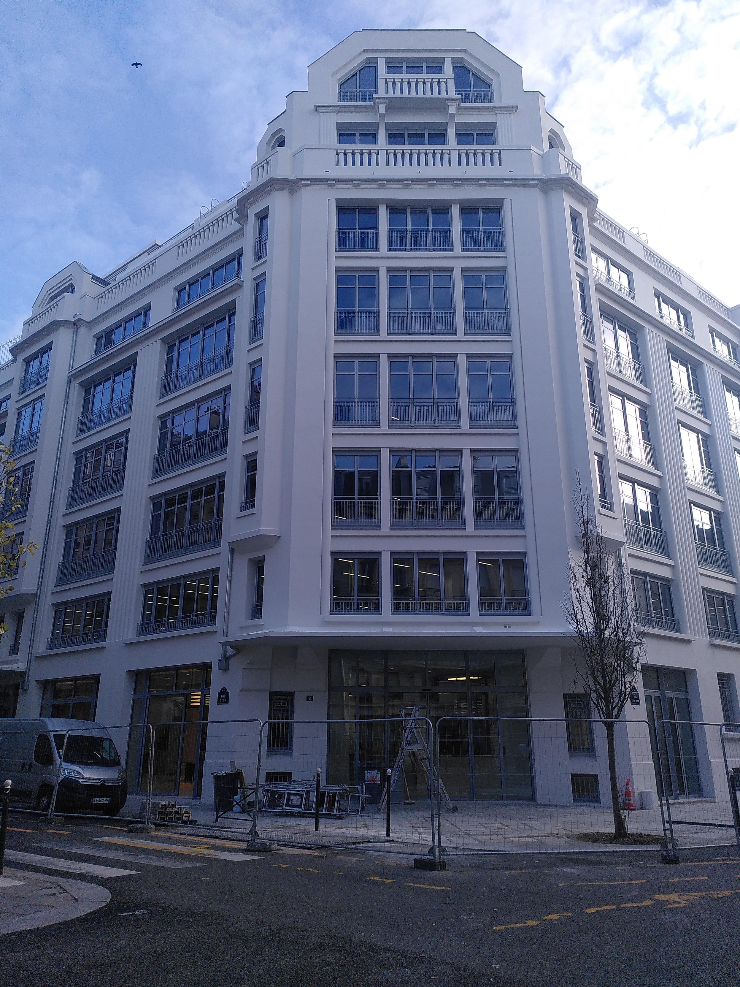 An office building in paris. It 8 stories tall with off-white wall, lot of tall windows all with a grey fence going up one third of each window. It's similar to common haussmanian style but less refined