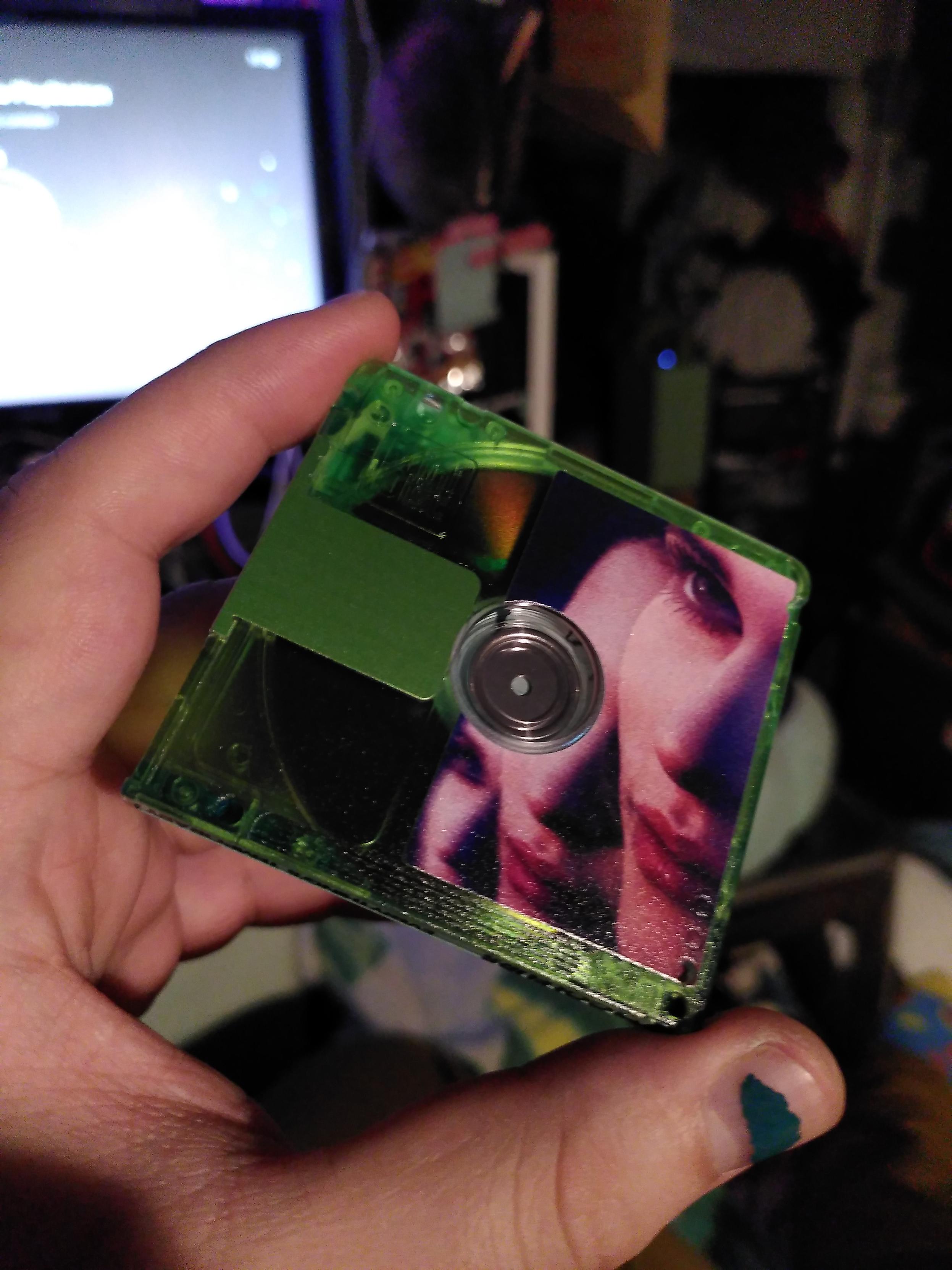 The other side of the coleurs' Everything's temporary minidisc. It's translucent green with the right side printed except where the mecanism to spin the disc is. The artwork depict a femme presenting face being repeated 3 time forming a diagonal
