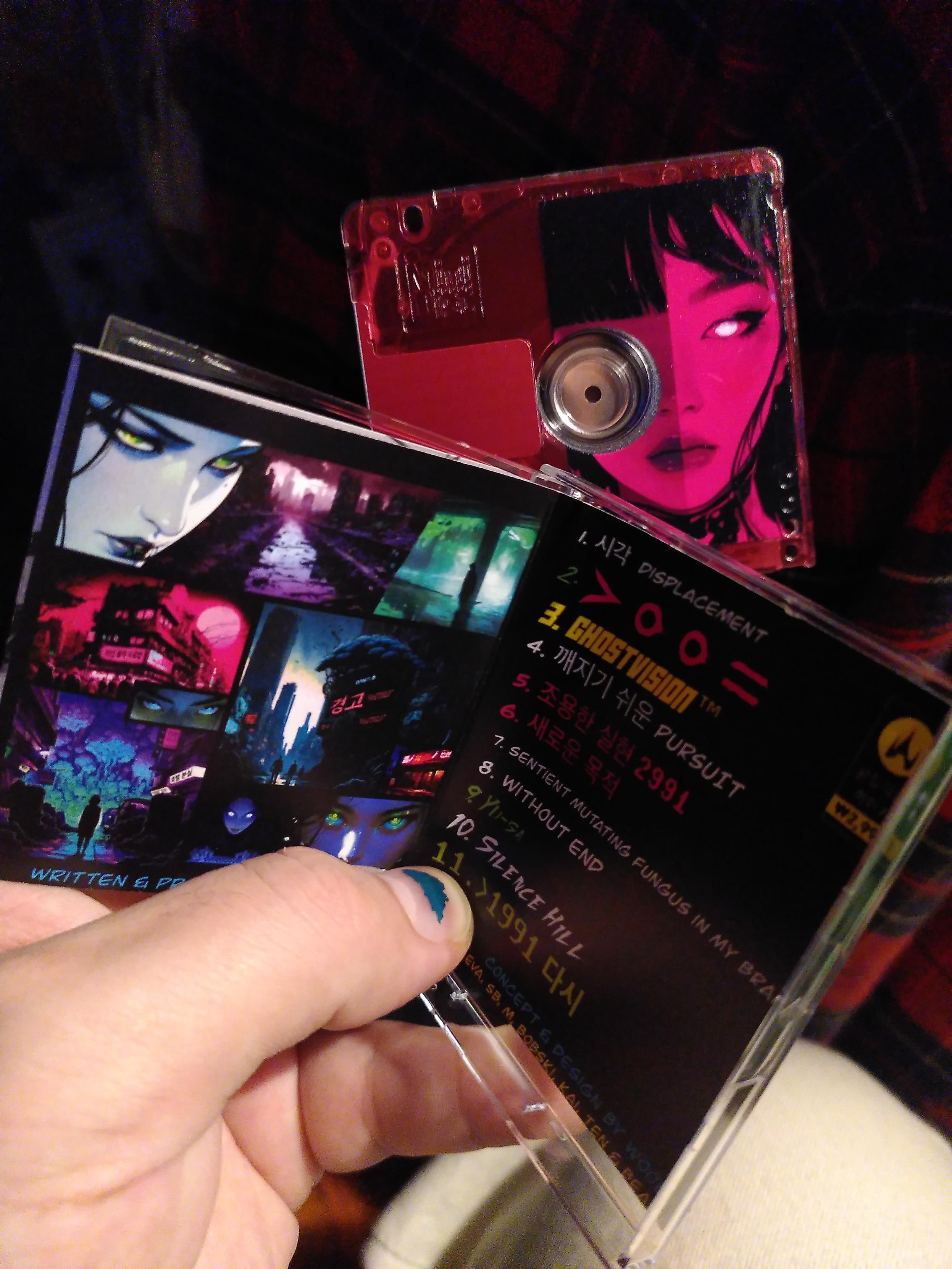 Me holding a red translucent minidisc and the case with the jacket open. There are multiple feminine faces, dystopian and brightly colored landscapes, the song list in multiple color and languages (english, korean? and symbols?)