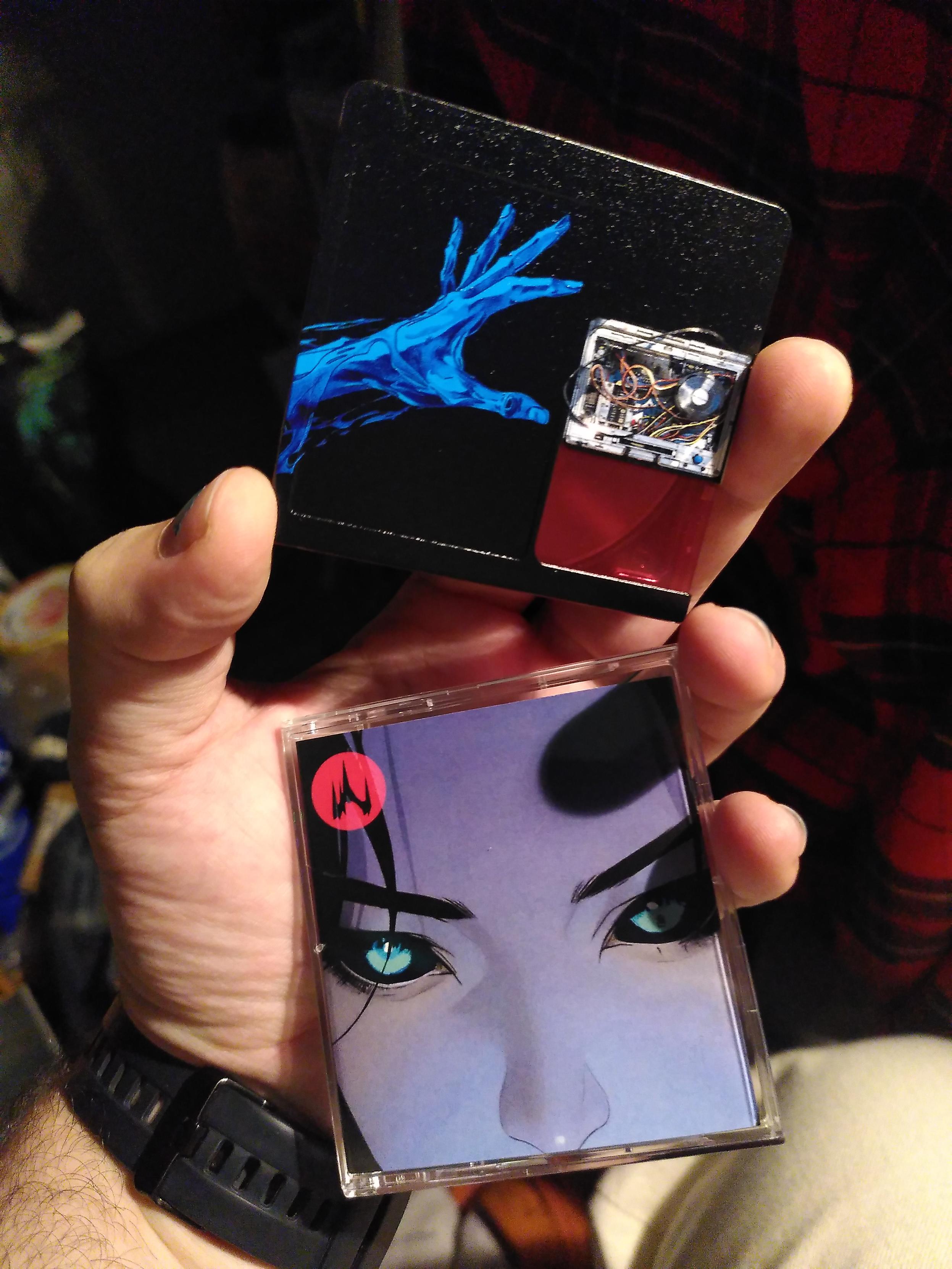Me holding the other face of the minidisc fully printed with a robot hand on black background and the front of the case, sporting the album cover