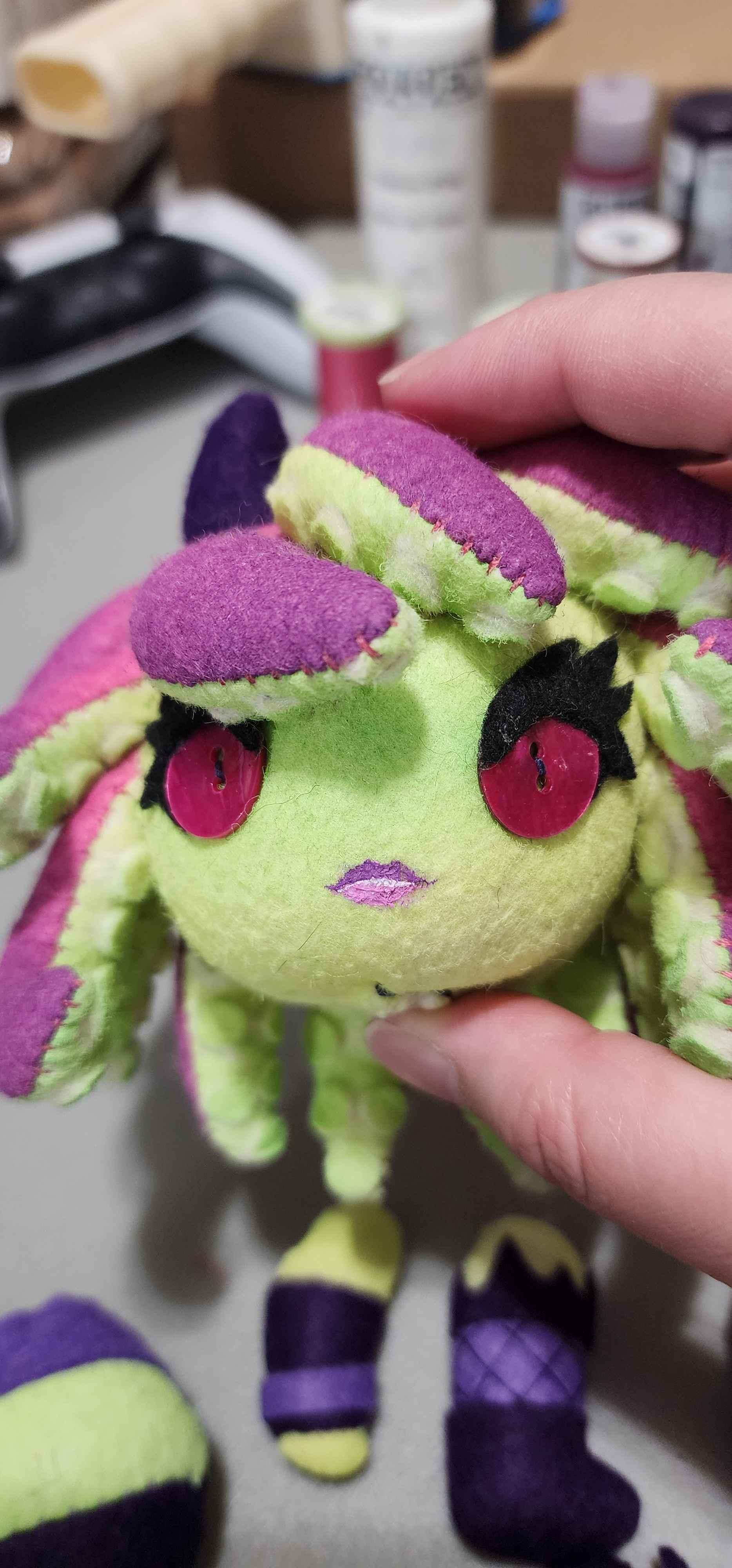 Head of a plushie held by 2 fingers. She has tentacles for hair, upper side purple and lower side green, green skin, eyes made of buttons with big black eyelashes and a tiny pink lipstick mouth