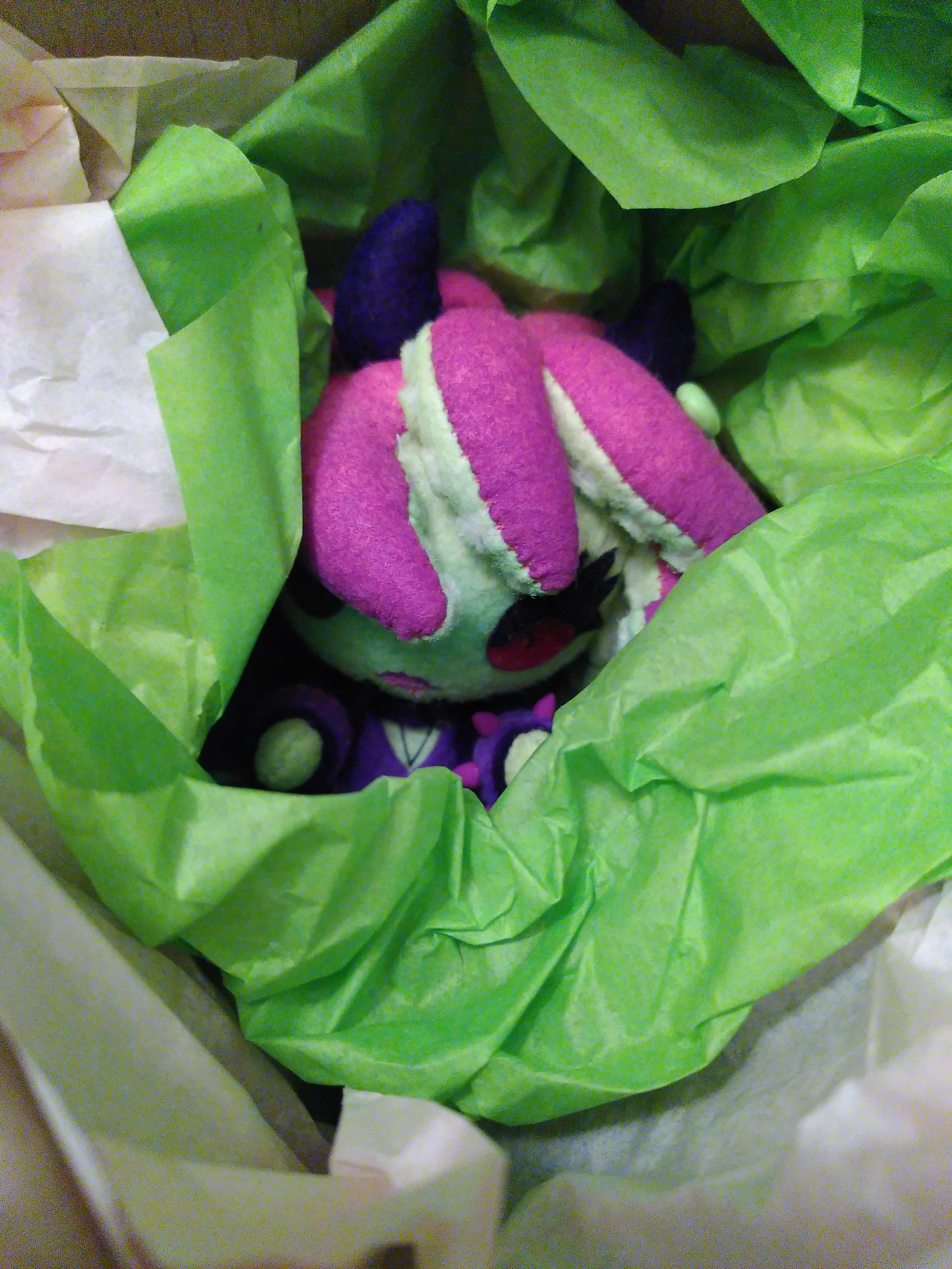 A small plushie cradled within soft green paper in a box. It have shiny redish pink buttons for eyes, green skin and pink tentacles for hair