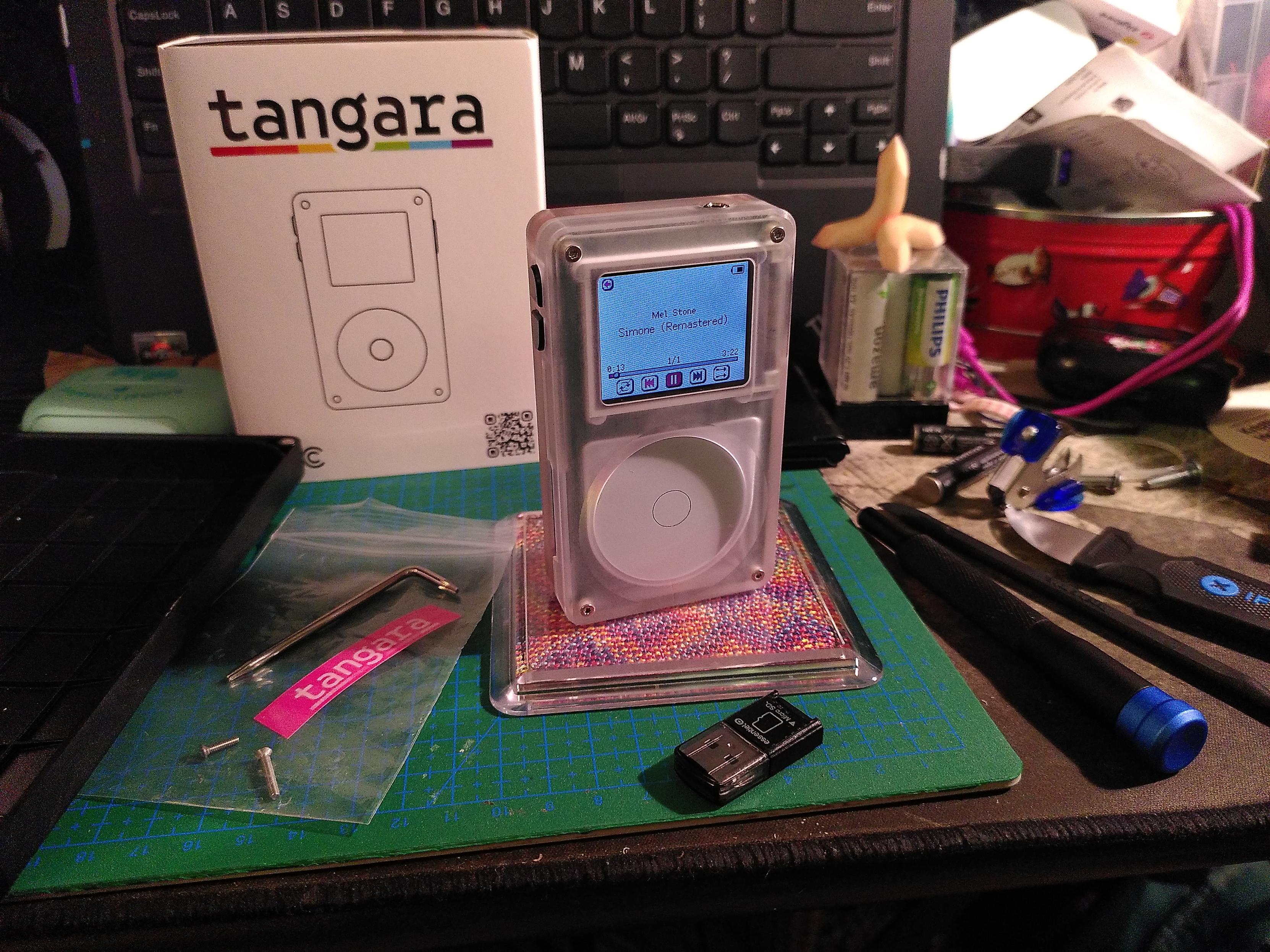 A tangara, cool tech zone's open source music player, on a busy desk. The device looks like an ipod with a clear shell, white circular input area and a low resolution color screen. It's standing upright on a plastic coaster containing some hand dyed fiber from https://www.fibraquarelle.com/. On the right are a few ifixit toools. On the left is a clear plastic pouch containing a tangara pink sticker, a tool to open the device and a couple of extra screw. In the back is the tangara box, with a lineart of the device and the title underlined by a rainbow. In front is a micro-SD to usb adapter.