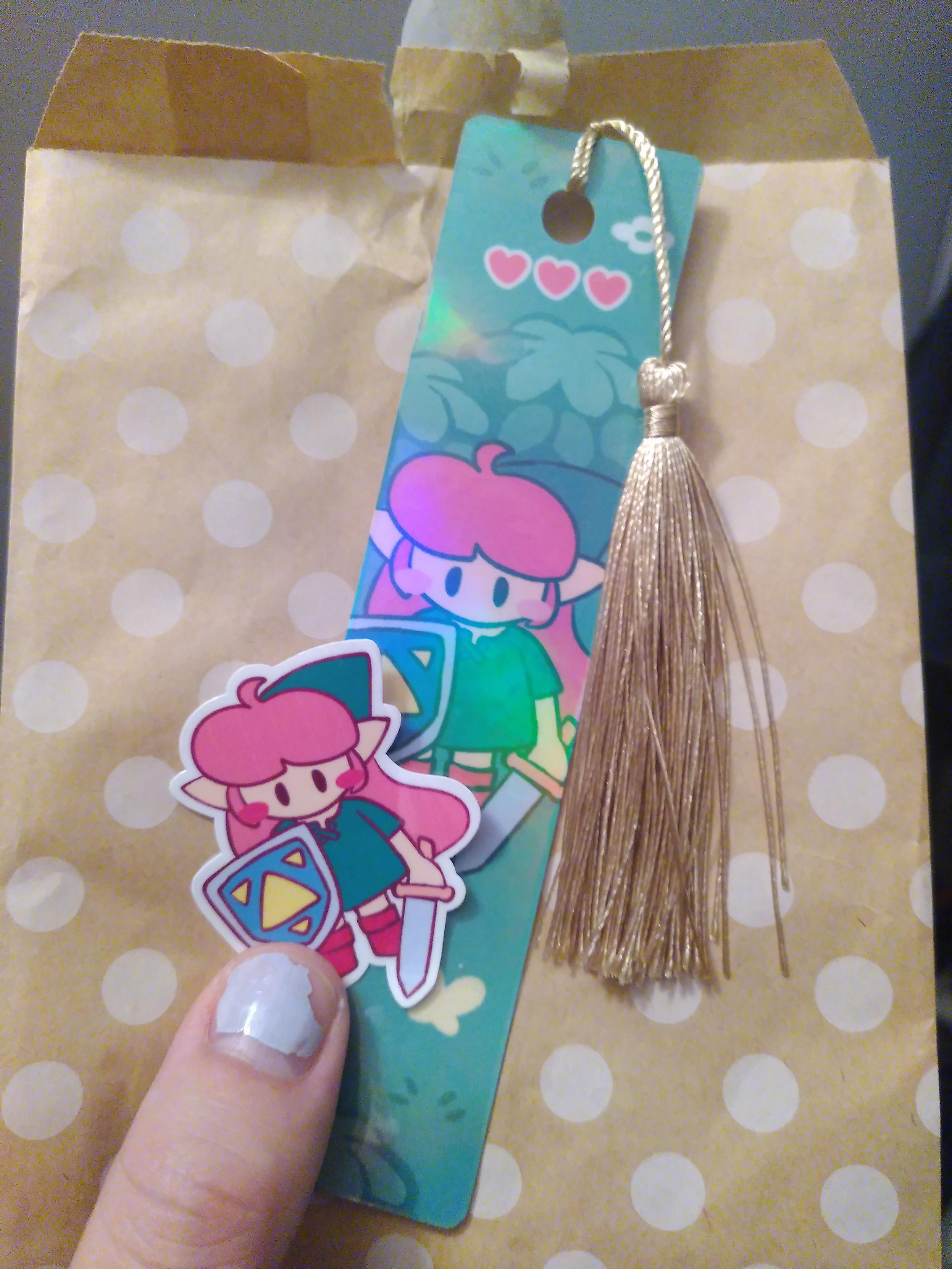A bookmark featuring the legendary girl and a bonus sticker with the same design. The bookmark is a long, thin piece of heavy paper with a hole at the top to let a golden string with a pompom go through.