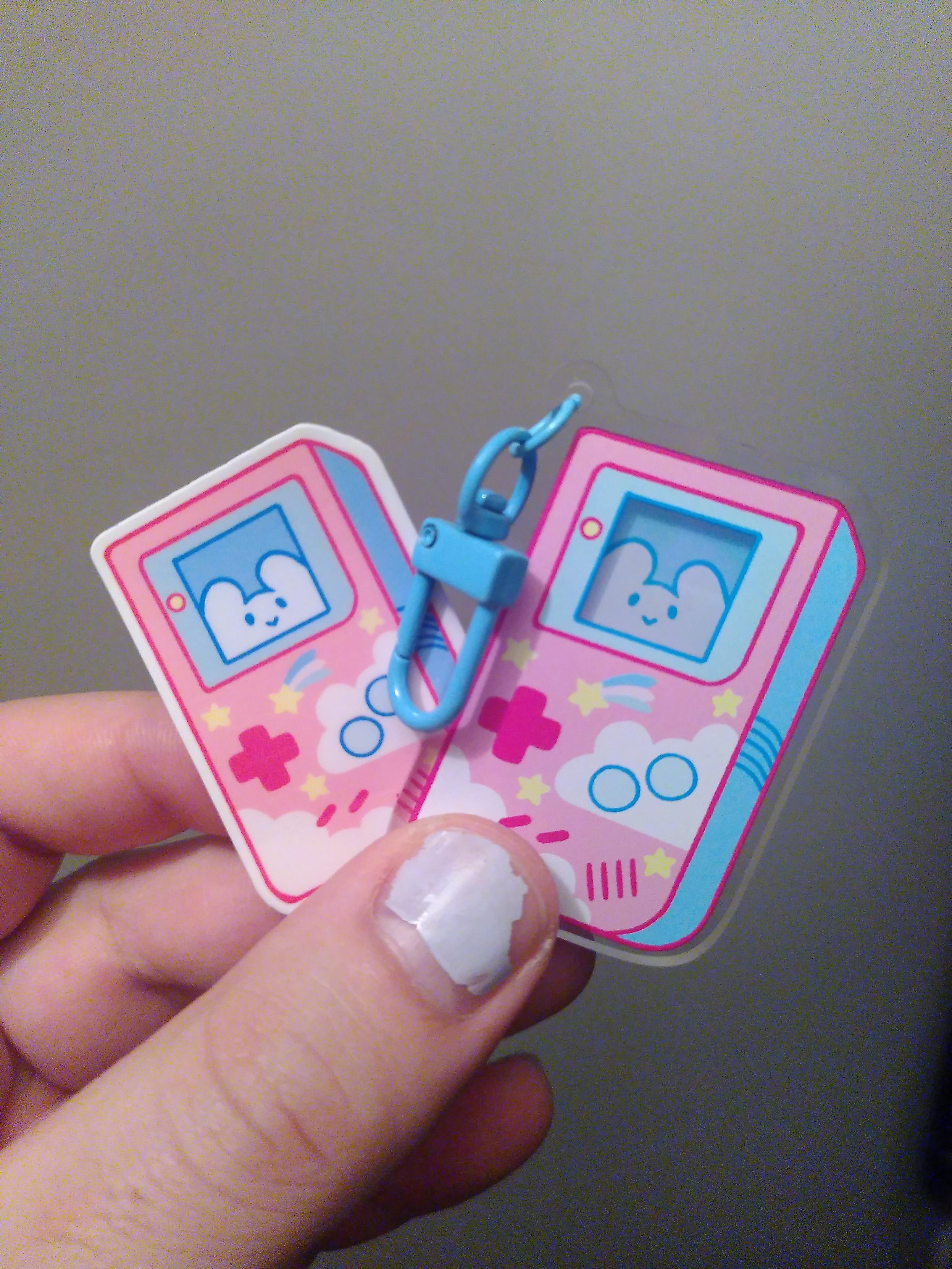 A strap acrylic charm looking like a cloudy pink gameboy with a jolly fellow behind the screen. It come with a sticker of the same design. Not pictured, the other side look like the back of a gameboy