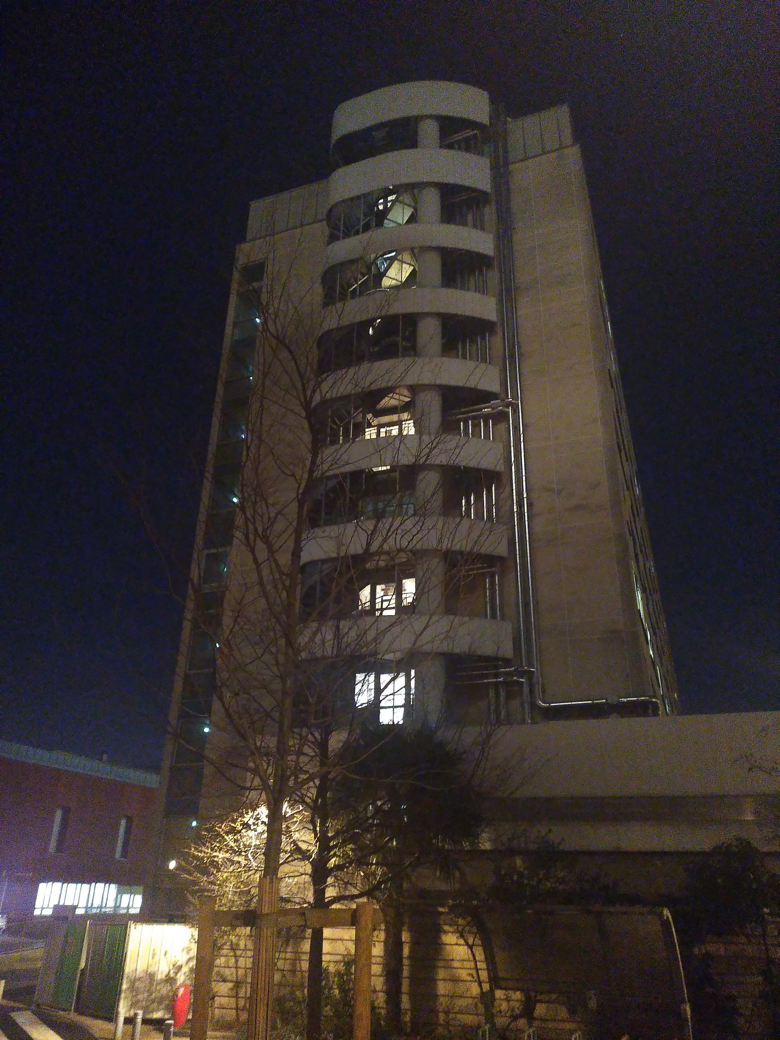 Picture of the side of a 8 stories high building by night. Each level have a round-ish balcony, each with a spiral stairway on the left side and large glass door, many with light. On the right of all the balconies is a set of metal pipe running all the height of the building