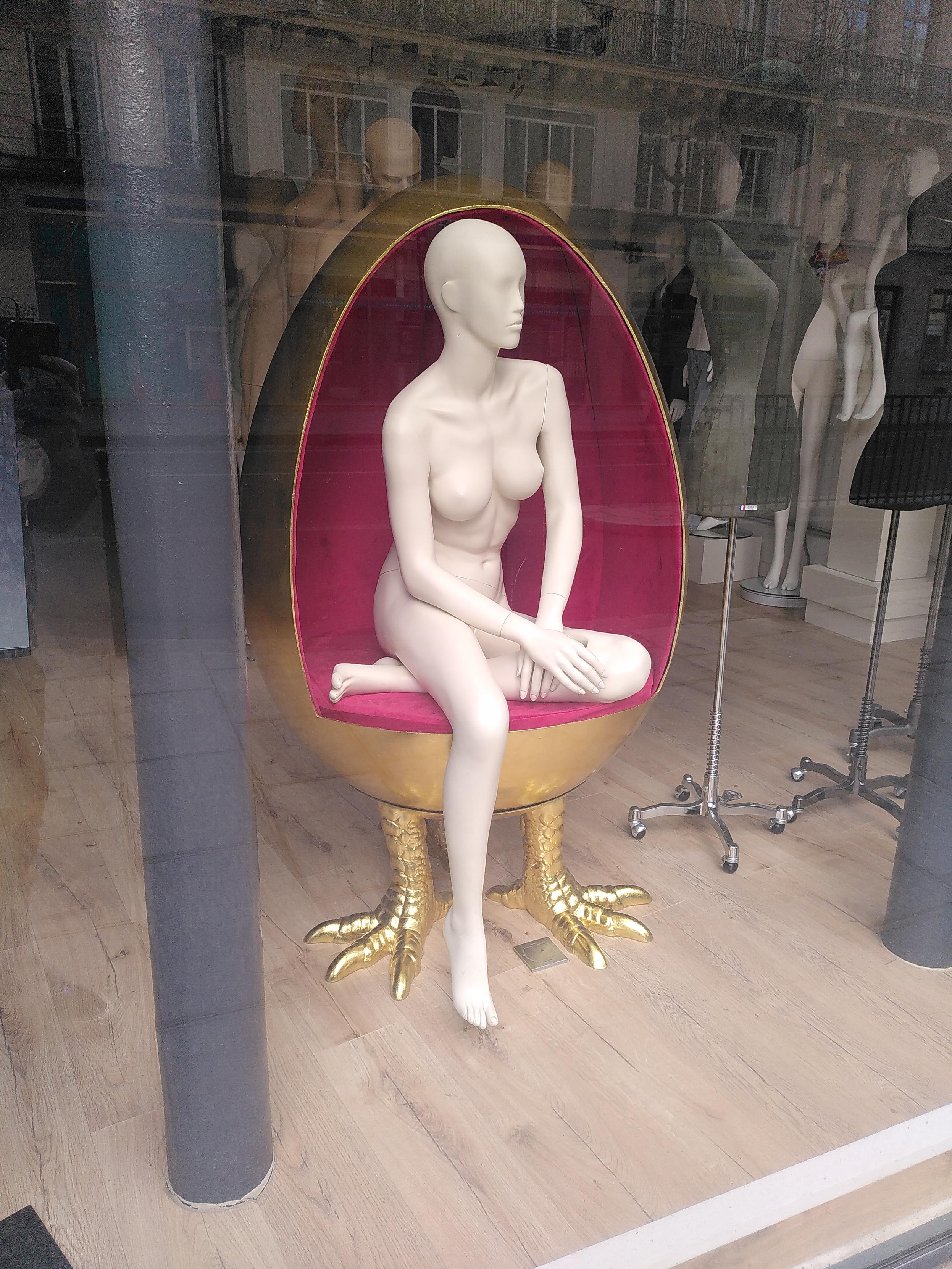 A mannequin sitting in a chair. The mannequin is white and featureless, posed sitting on one leg and the other dangling down. The chair is shaped like an egg with a pair of chicken legs, the inside is wrapped in red velvet and the outside is all gold 