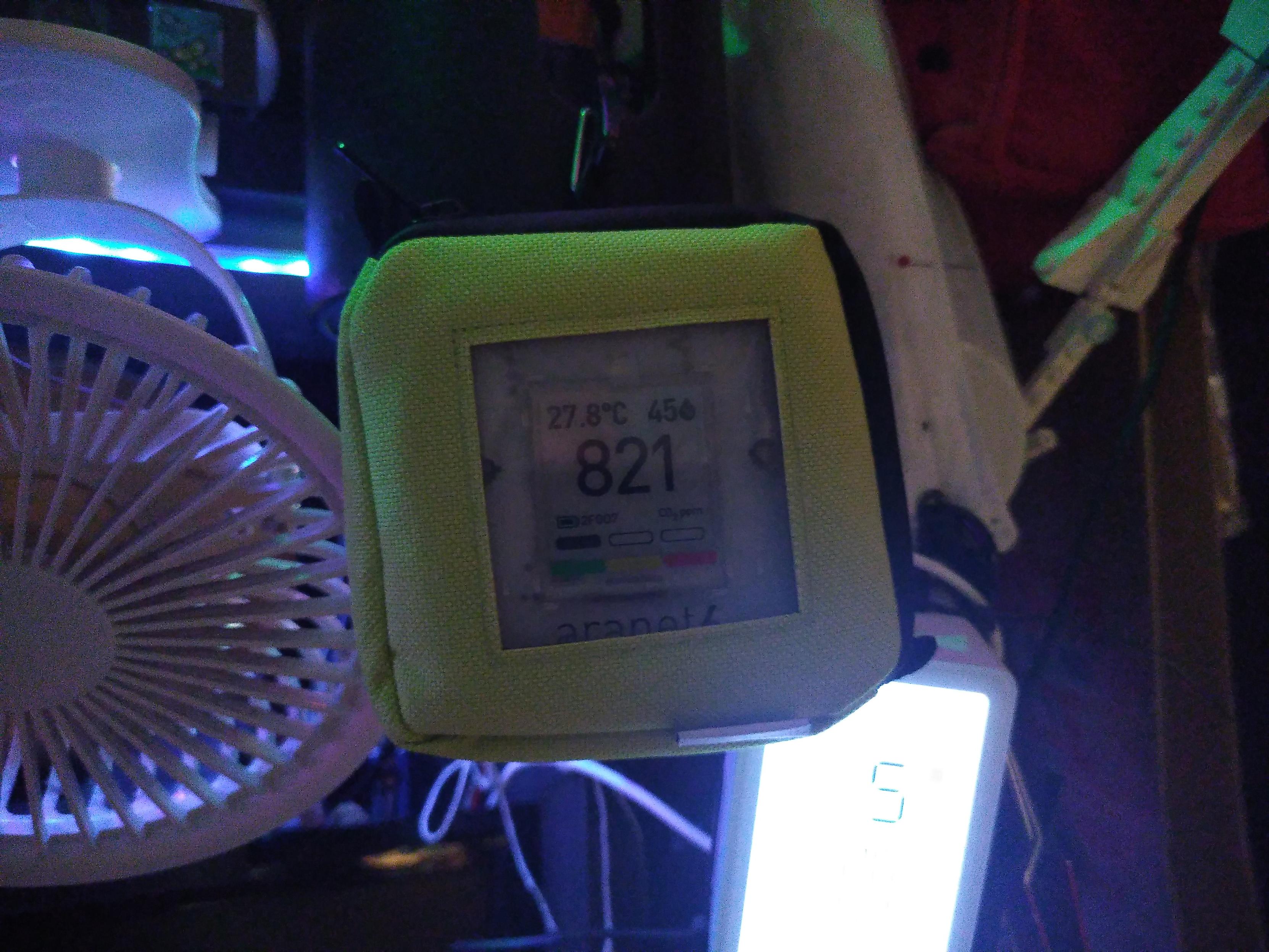 An aranet4 inside a pastel green case. On the left side is a small electric fan and below it is an ikea environment monitor too bright to be readable on the photo. The aranet4 is a small battery powered device that measure humidity, temperature and CO² level.