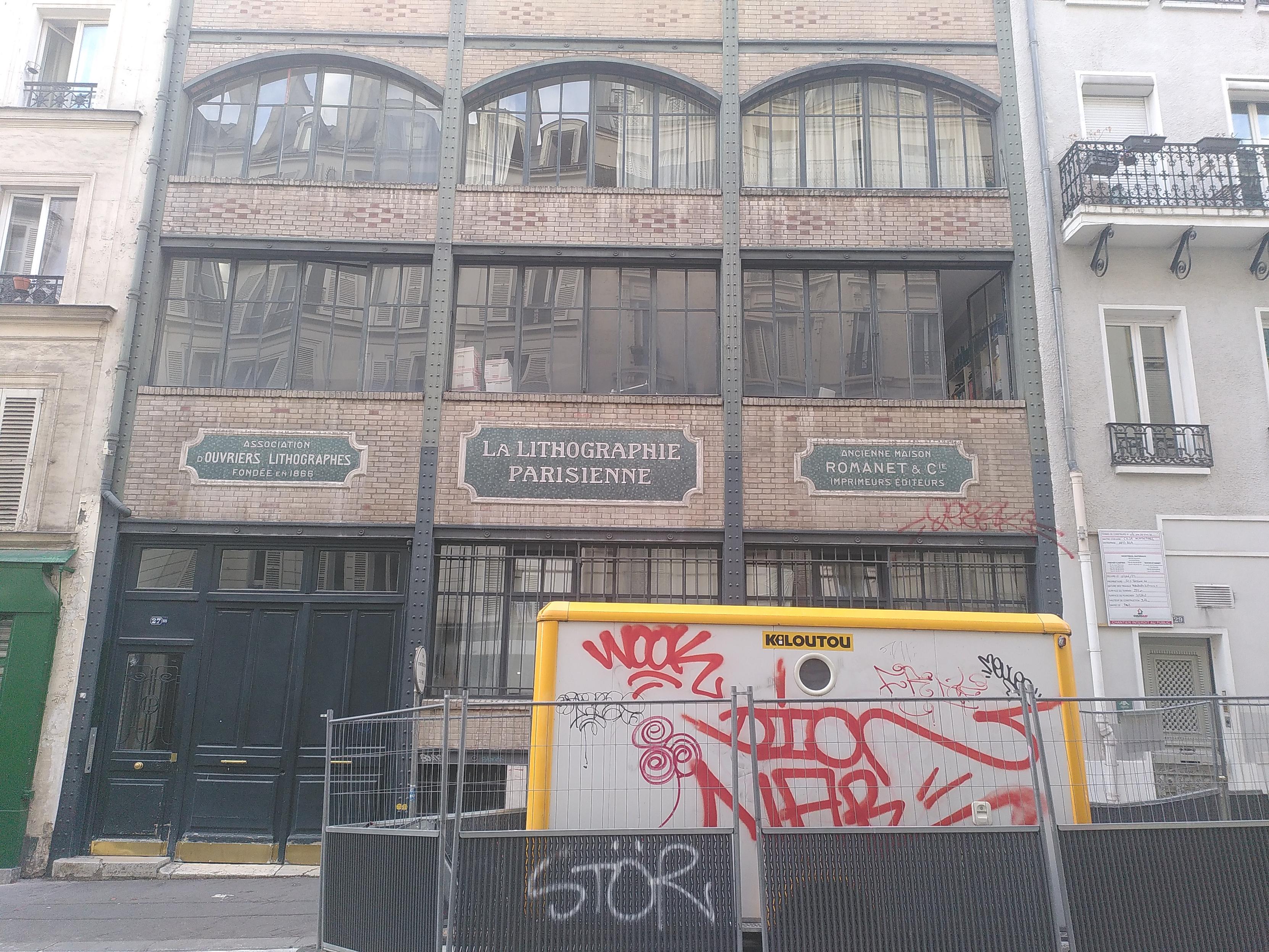 Facade of a building in paris. It's made of a steel structure in dark gray, brick in very light red plus a few dark red making some pattern and very wide window made of tall and narrow glass panel. On the second floor, on the brick are some large label, mentioning it was home of a business funded in 1866 doing lithography