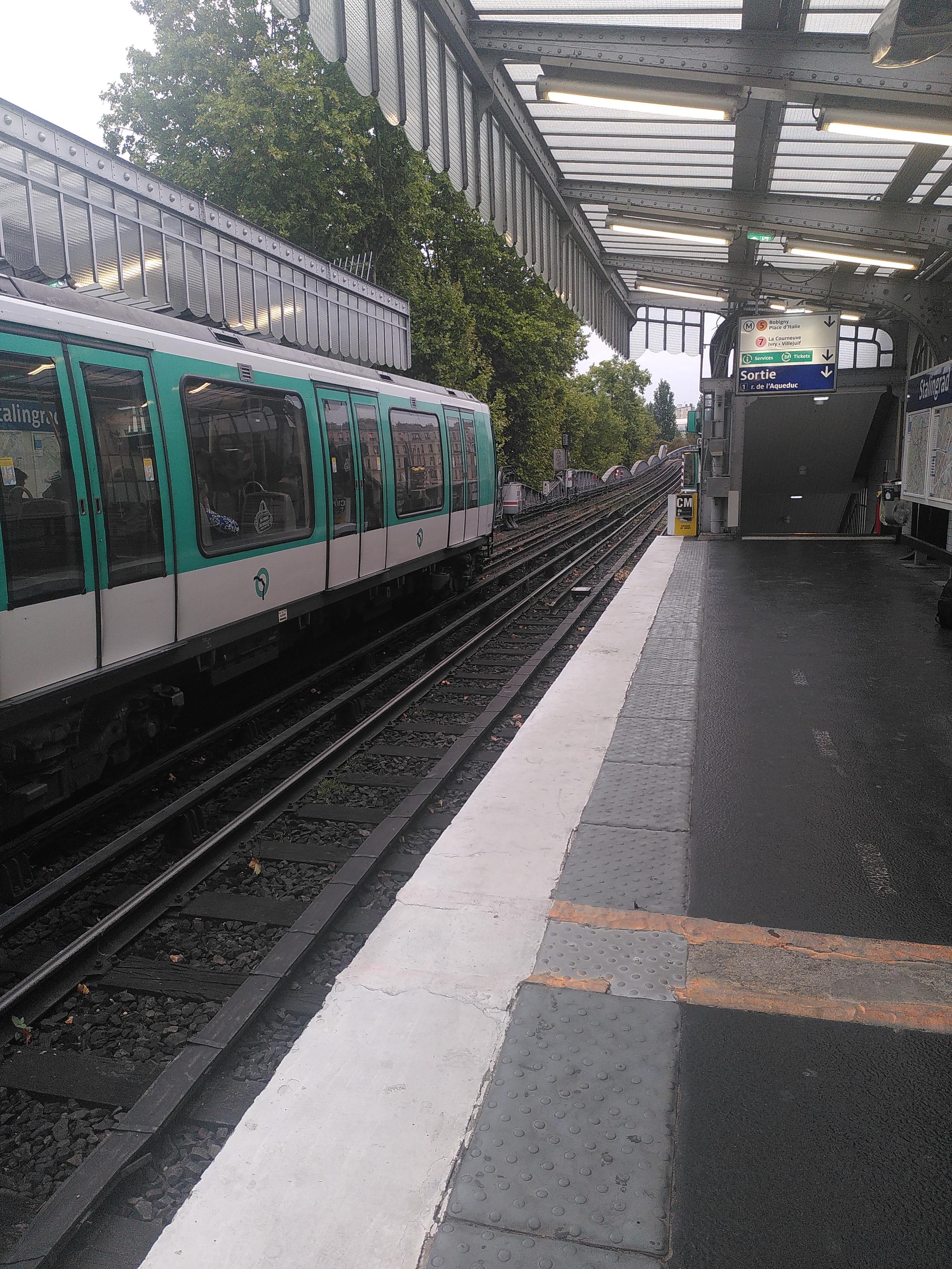 Pic of an outdoor metro station. The waiting area is covered witg glass and steel structure with lot of rivet. The ground is three colors, black away from the rails, a textured gray strip toward the rails the a white stripe. Behind the station is a bridge made of the same steel with rivet material, bordered by trees
