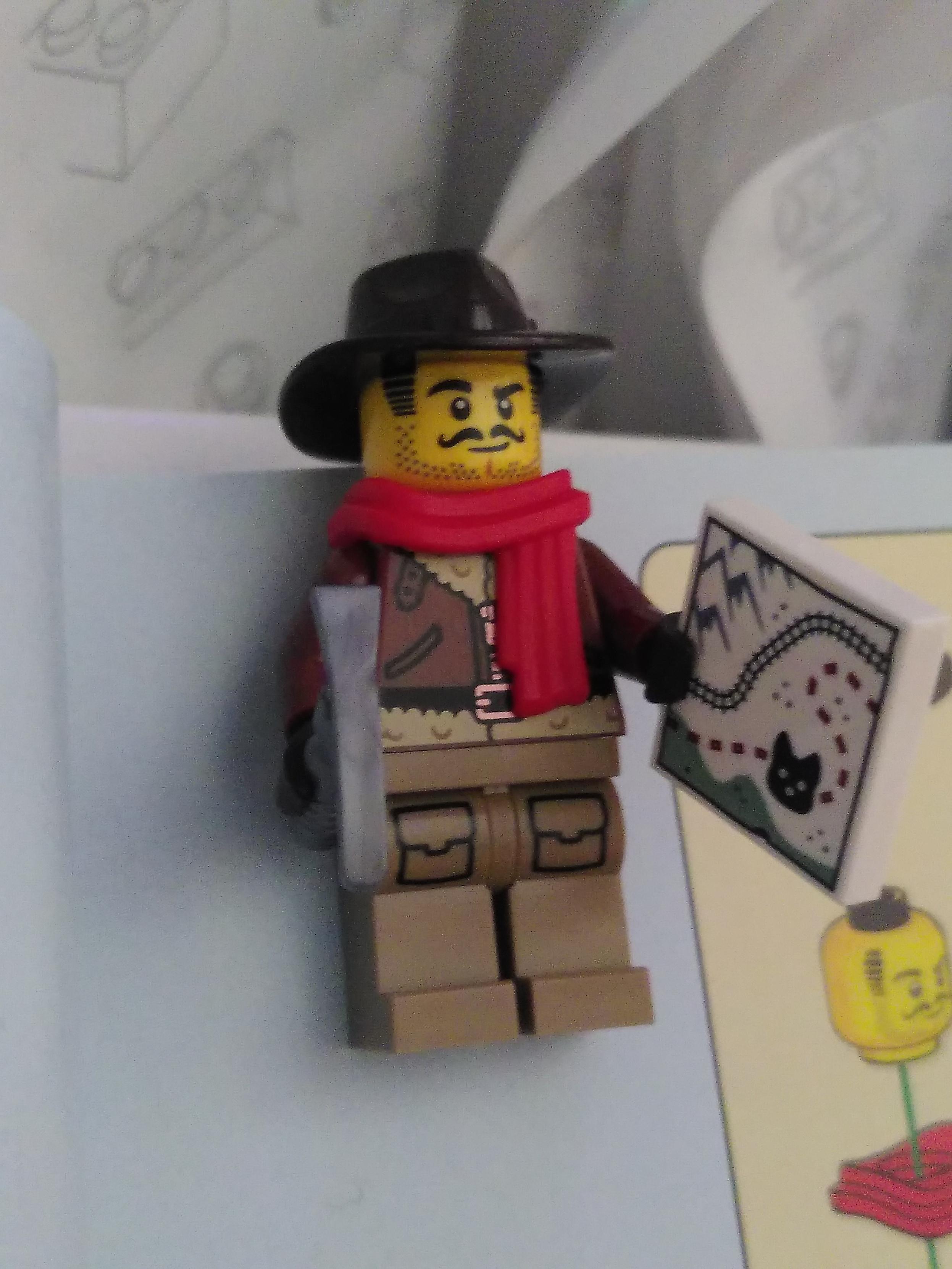 A lego mini figurine sporting a light brown pant with two (printed) front pocket, a (printed) leather jacket lined witg fur, an impressive mustache and a two day beard, a leather explorer hat and a red scarf. It's holding in its right hand a pickaxe and in its left hand a map, mostly covered in snow, with mountain in the top left corner, a bit of green on the bottom side, train track going from middle left to upper right, a red dashed line from lower left side to upper right side doing an S shape with a black icon on the dashed red line looking a but like a neocat or a husky head