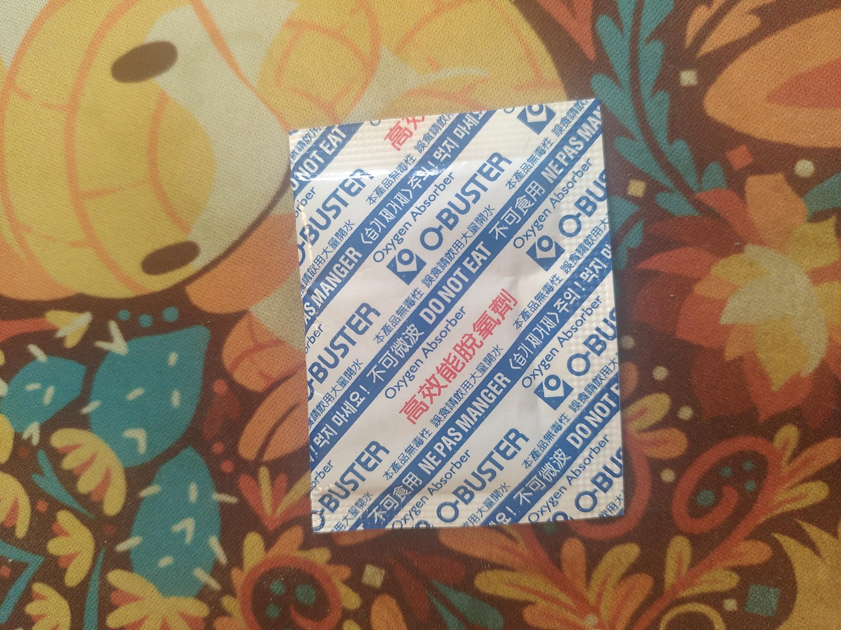 A packet of oxygen absorber. It's covered in pattern of white line and blue lines. The white line are covered with oxygen absorber and o-buster written in blue and some east asian red character. The blue line are coveres with do not eat, ne pas manger and three other sentences in east asian character (probably korean, chinese and japanese).