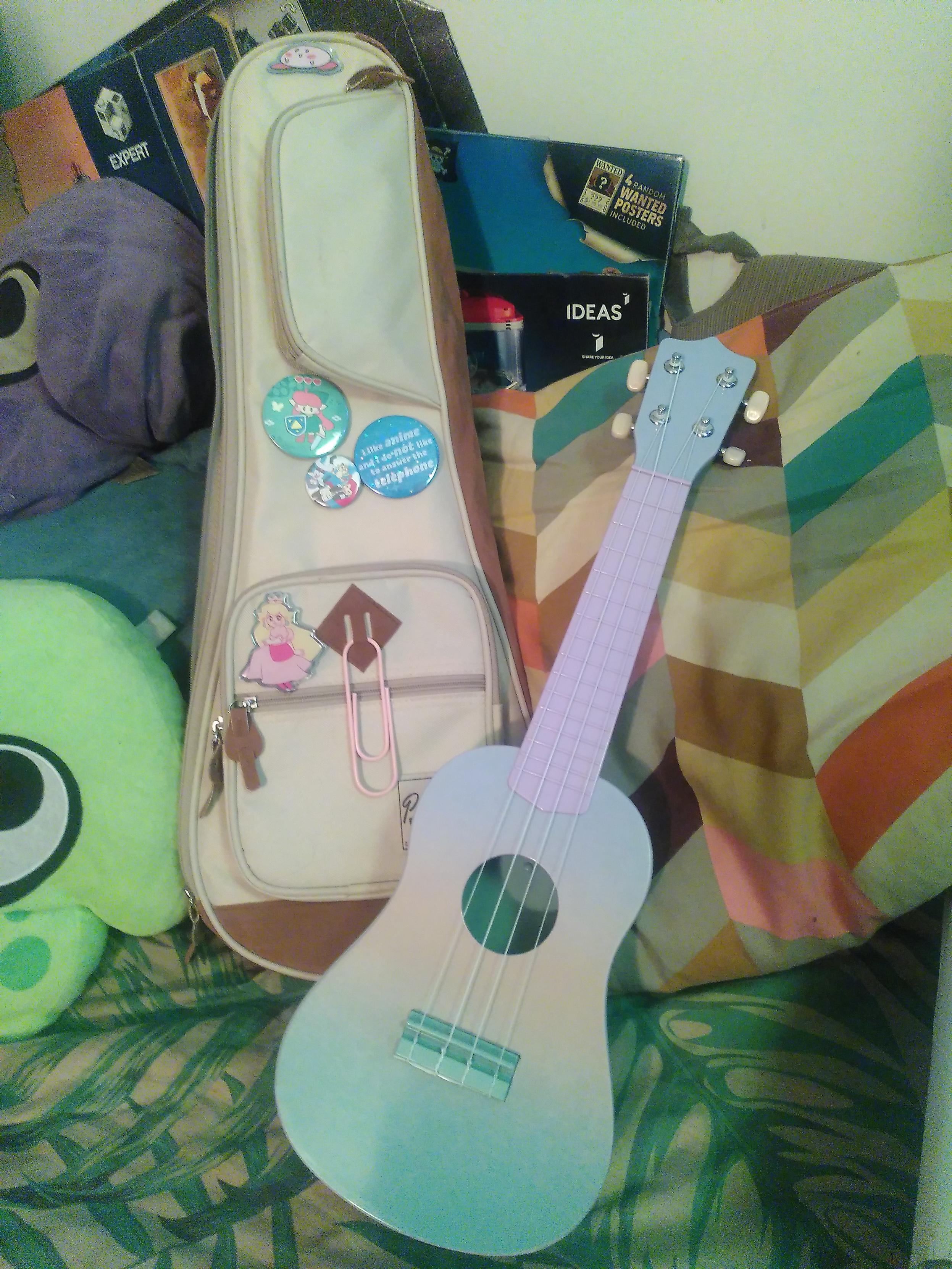 Photo of a powerpad yukulele bag and a cheap plastic yukulele. Bag is mostly very light brown with mid brown accent for the handle, the shoulder strap (not visible), a small panel at the bottom and the sides which might touch the ground. It's decorated with a bunch of buttons and pins from doodlemancy. The yukulele is made of plastic, colored in a pastel gradient starting green at the bottom, going a very soft orange then purple, pink for most of the fret and purple again for the head. It can be accorded and play ok, at least to my ear. In the background is a blahaj plushie, two splatoon squid plushie in purple and neon green, an ikea cushion
