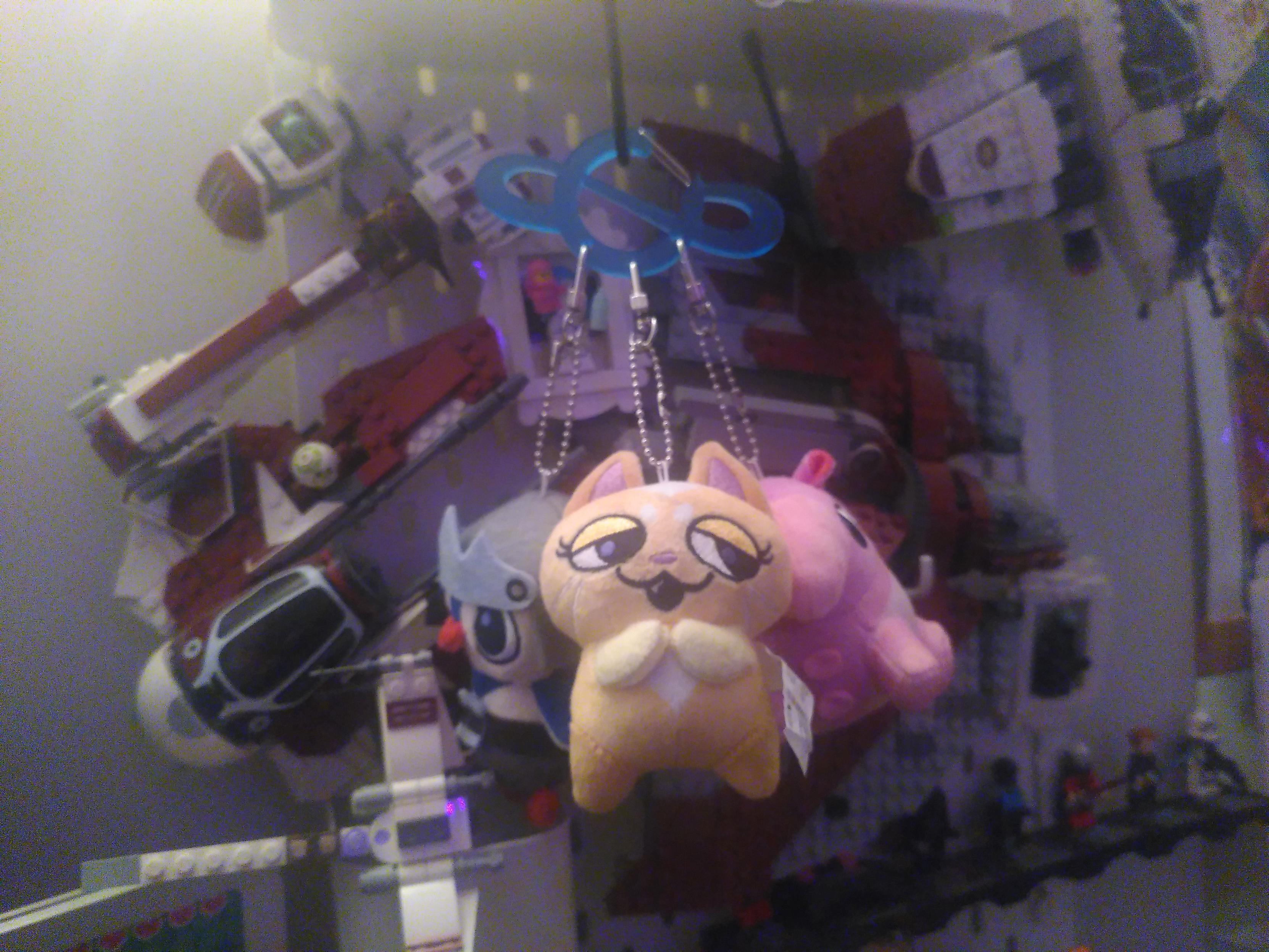 Photo of 3 keychain plushie attached to a carabiner. The plushie are from the show Knights of Guinevere and are the space trio. Therd is a yellow cat, a bright pink tapir and a penguin in knight armor. The carabiner is translucent blue acrylic in the shape of the planet with a wavy ring, I think the logo of the show. Behind out of focus are a bunch of lego star wars attached to a peg board