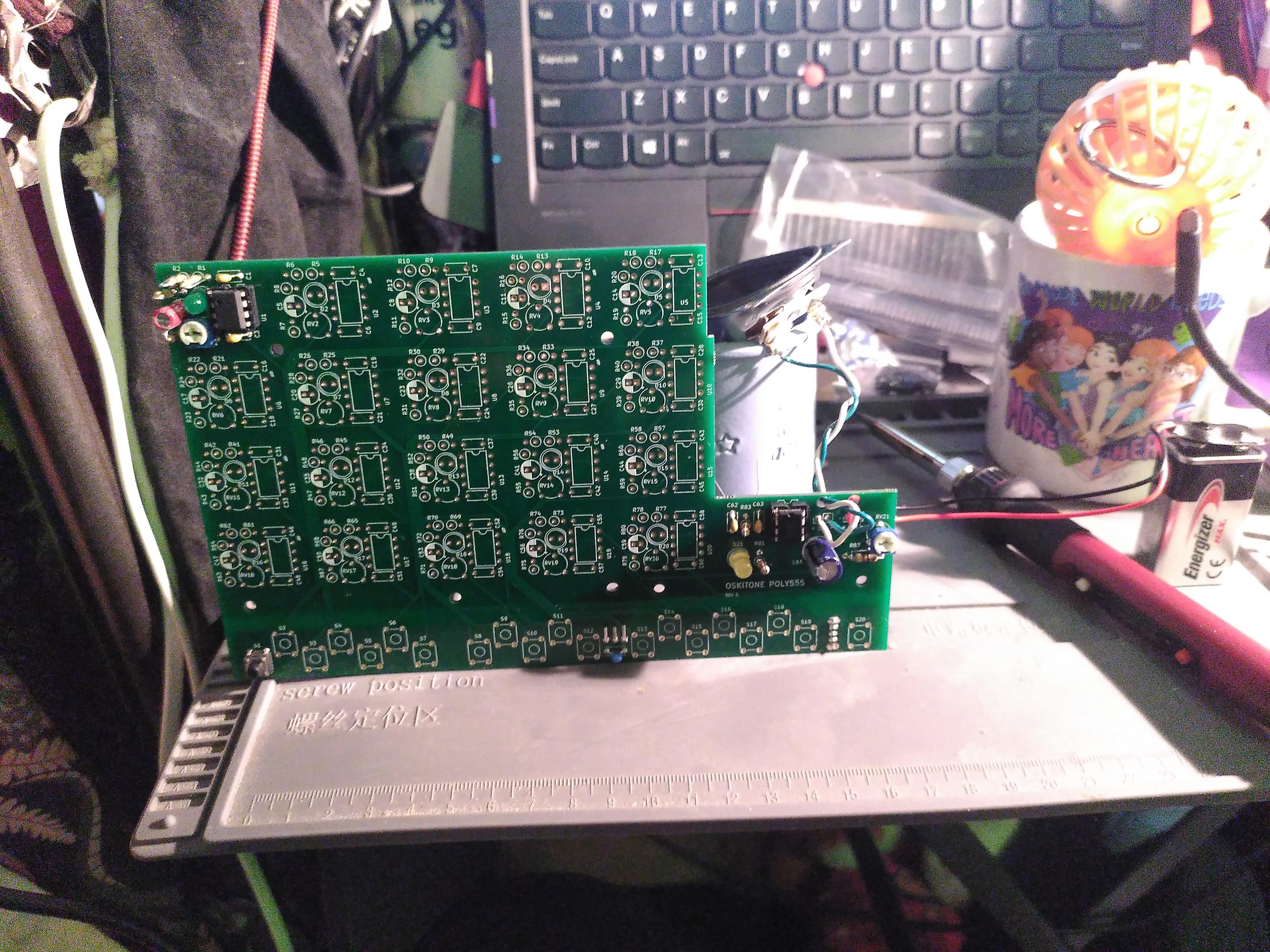 Photo of a pcb standing upright. Only a small part have been populated with through-hole components. On the right is an amp circuit, a led to indicate when power is on, a 9v battery, a speaker and a pin from two components hidine underside, a switch and a power converter. On the top left is a circuit built around a 555 timer and a led and in the bottom left a momentary switch to power the noise making circuit. There are 19 circuit and switch unpopulated. In the end, it will make a poly555, a polyphonique analogique synthetizer