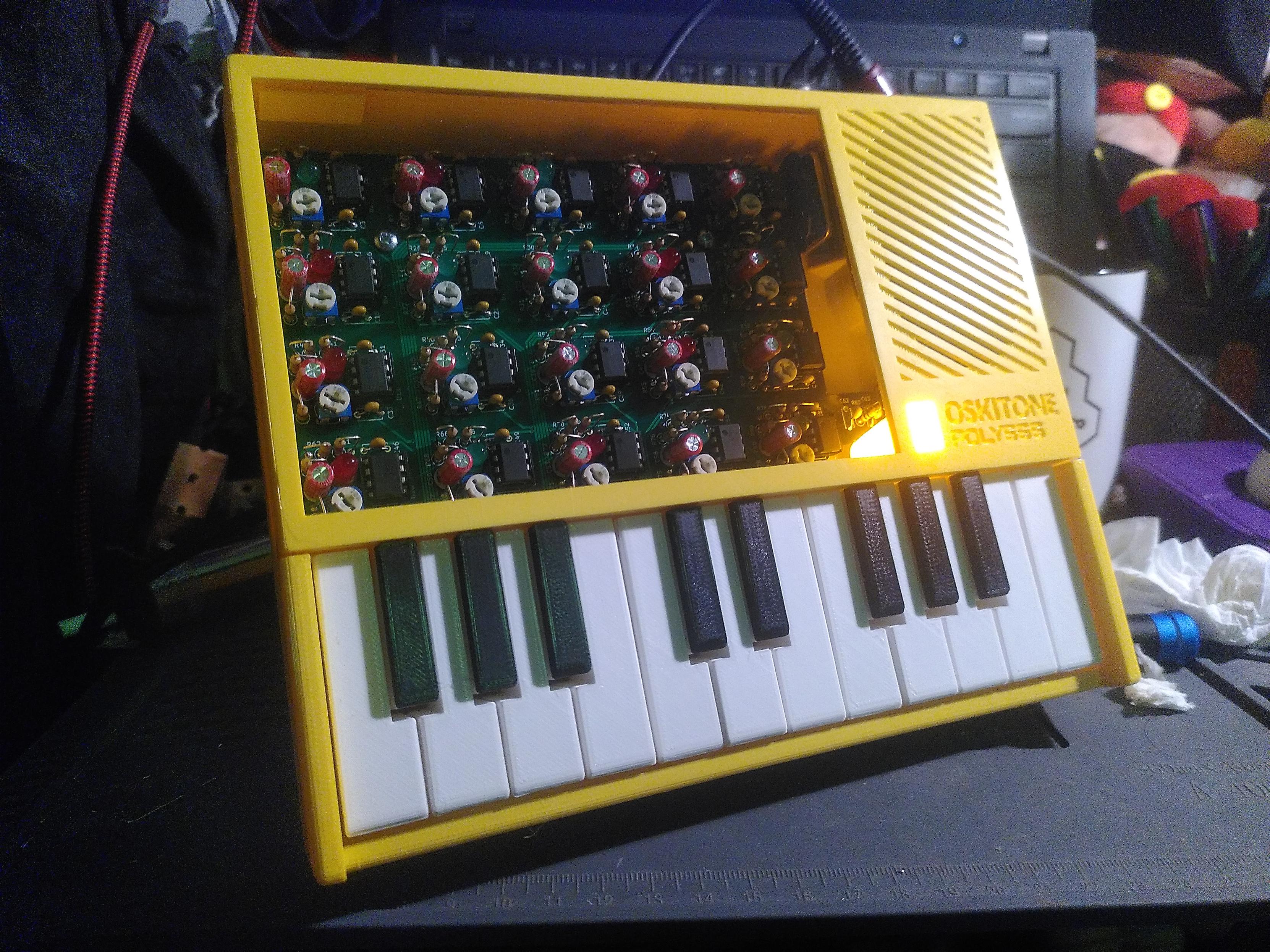 Photo of a yellow poly555, an analog polyphonique synthesizer. Visually it's a rectangle 16cm high and 20cm wide with 3 distinct area. The bottom part have 20 keys with white and black, like a piano. The upper left part which take a bit more than two third is a transparent panel exposing a pcb with 20 groups of component in a grid of 4 row and 5 columns. Each group is a 555 timer, producing a note when the matching key is pressed. Each timer also have a led, green for white keys and red for black keys. The upper right is a grid for the speaker and is part of the case. The case is yellow and 3D printed, giving it a texture specific to FDM
