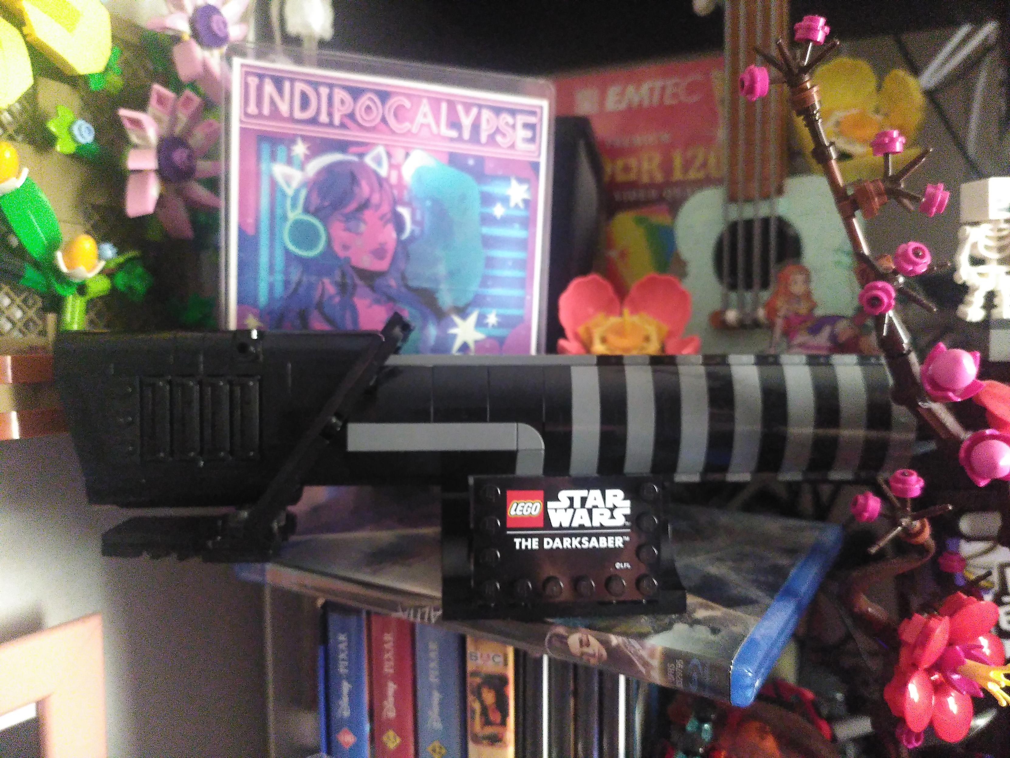 Photo of a lego lightsaber. Maybe 1:1 scale. Around it out of focus are a few DVD and blu-ray (half I haven't watched), lego flower on a treillis, lego yukulele and flower, lego plum blossom, a lego minecraft skeleton and an indipocalypse mini-poster featuring a cute lady with cat-girl headphone in pink and blue neon colors