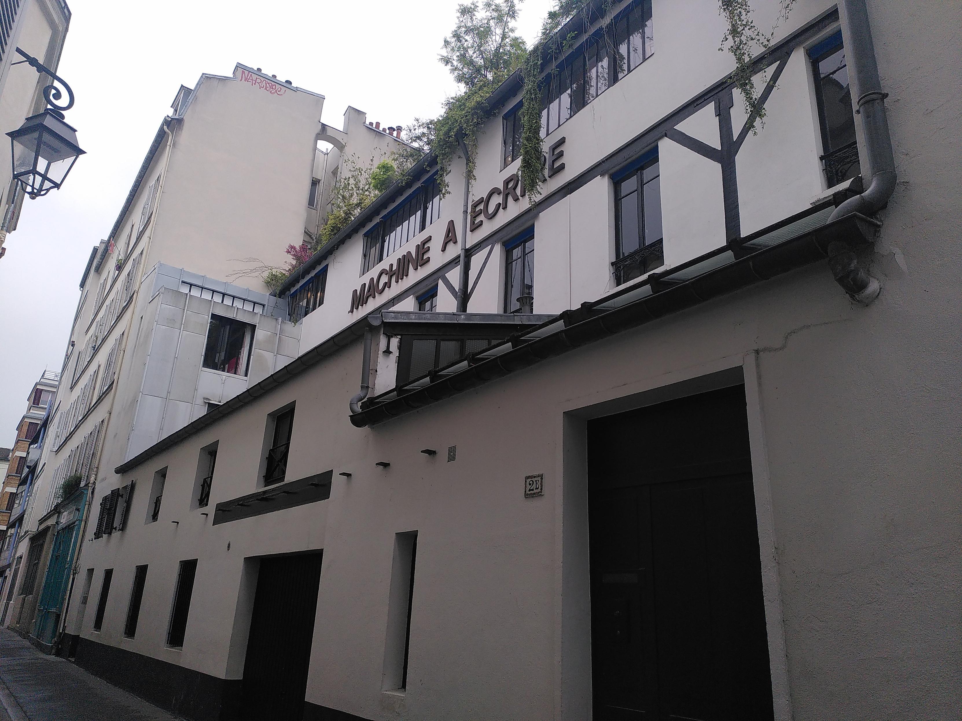 Photo of a small alley in paris, showing the side of an old factory, probably type writer as it's written on the side. Everything seems to have been restored not too long ago while still exposing some underlying element. On top of it is some greenery going over the side