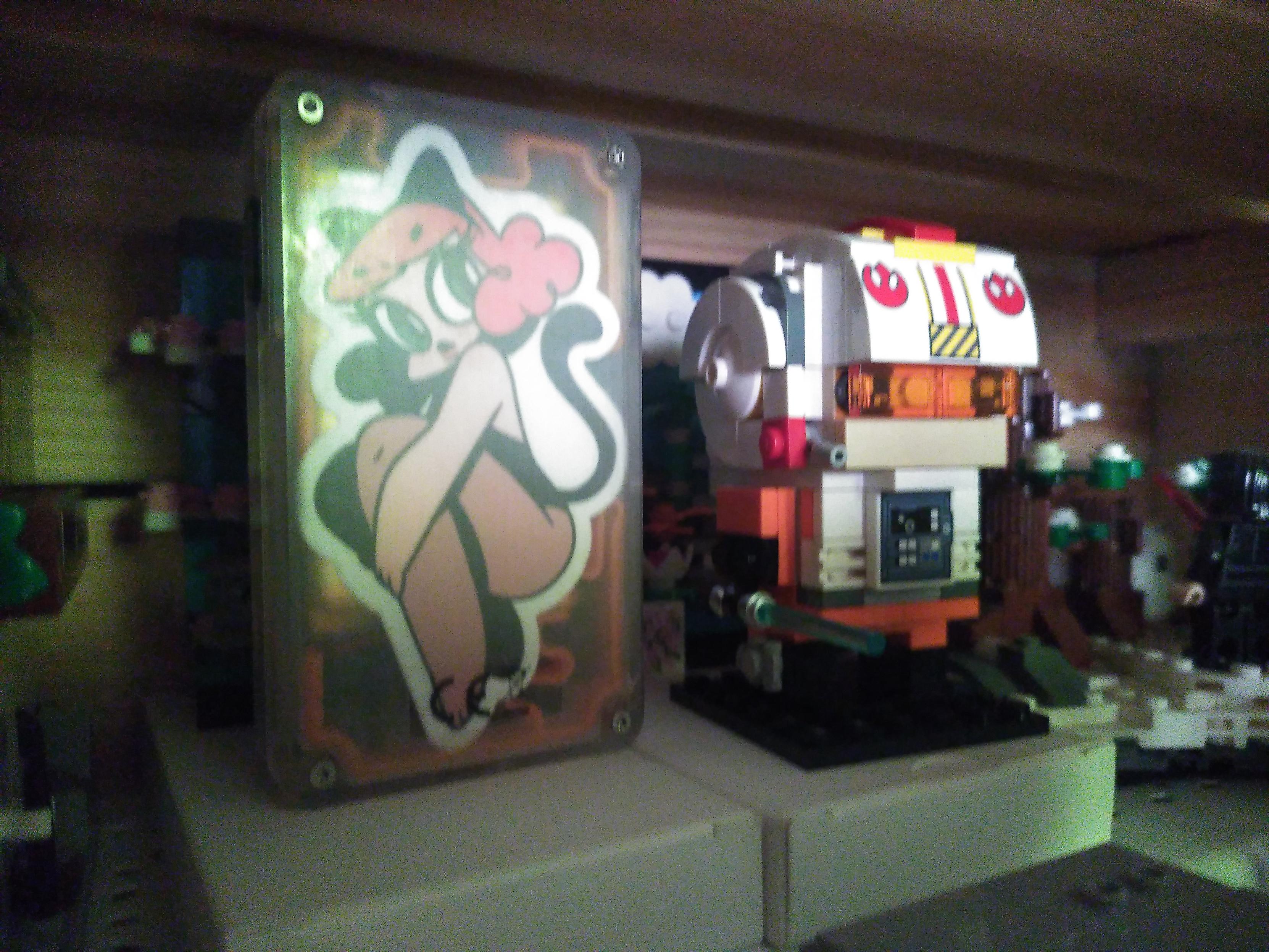 Photo of a tangara music player and a lego brick headz of luke in x-wing pilot outfit. The lighting isn't great. Only the back of the tangara is visible, made of translucent plastic showing inside an orange 3D printed contraption holding the battery in place and a sticker stuck inside. The sticker depict a drawing of a lady with cat ear and tail, curvy red hair, bar arms, black body and stockings covering her legs. The lego brick heads is luke in his bright orange andnwhite outfit, with a white helmet, translucent orange visor and rebel logo. He is holding a blue lightsaber and might have a blaster strapped to his tiny leg