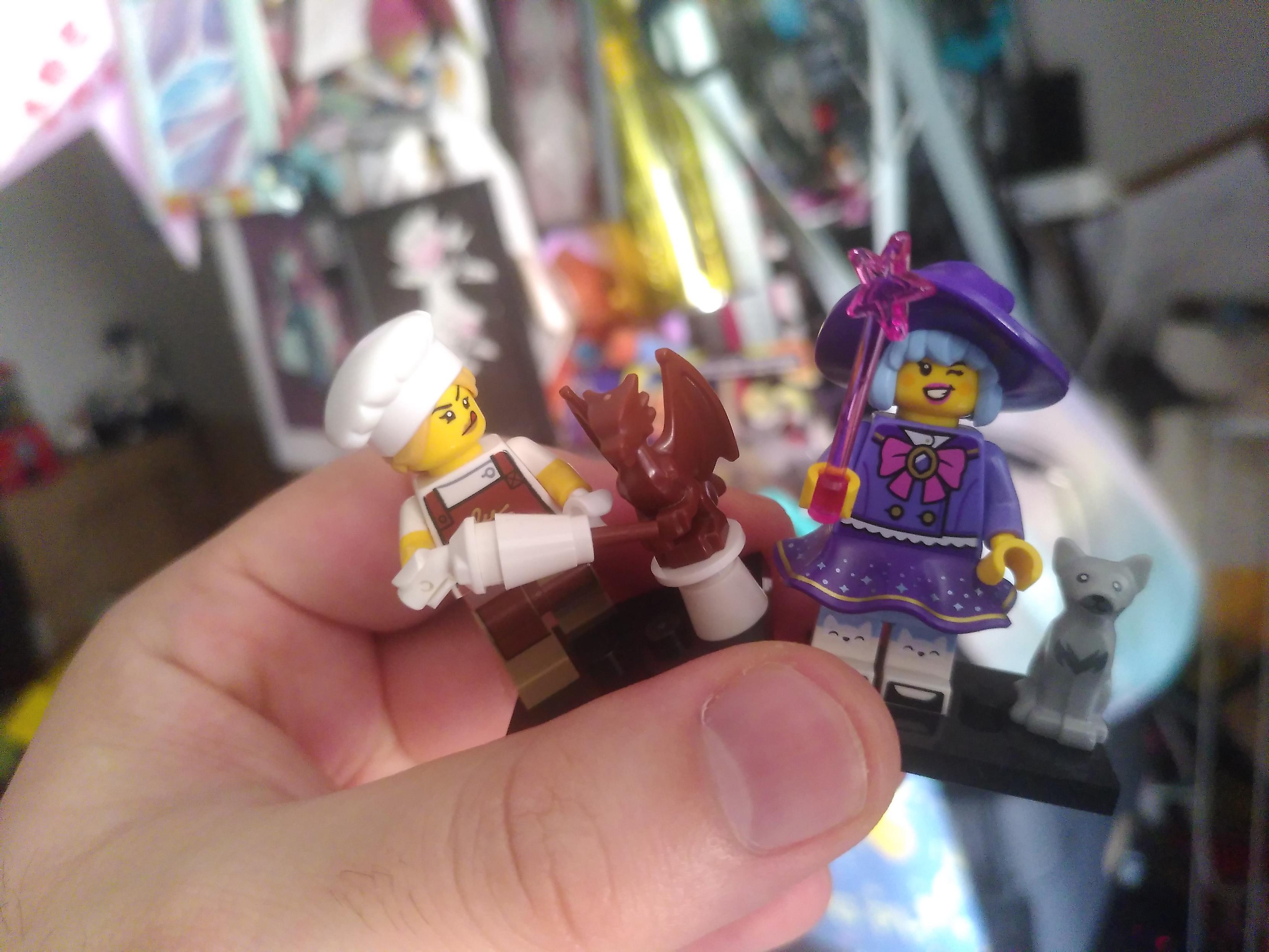 Photo of a hand holding two minifigurine. One is a lady cook making a chocolate dragon, one is a cute witch with a gray cat
