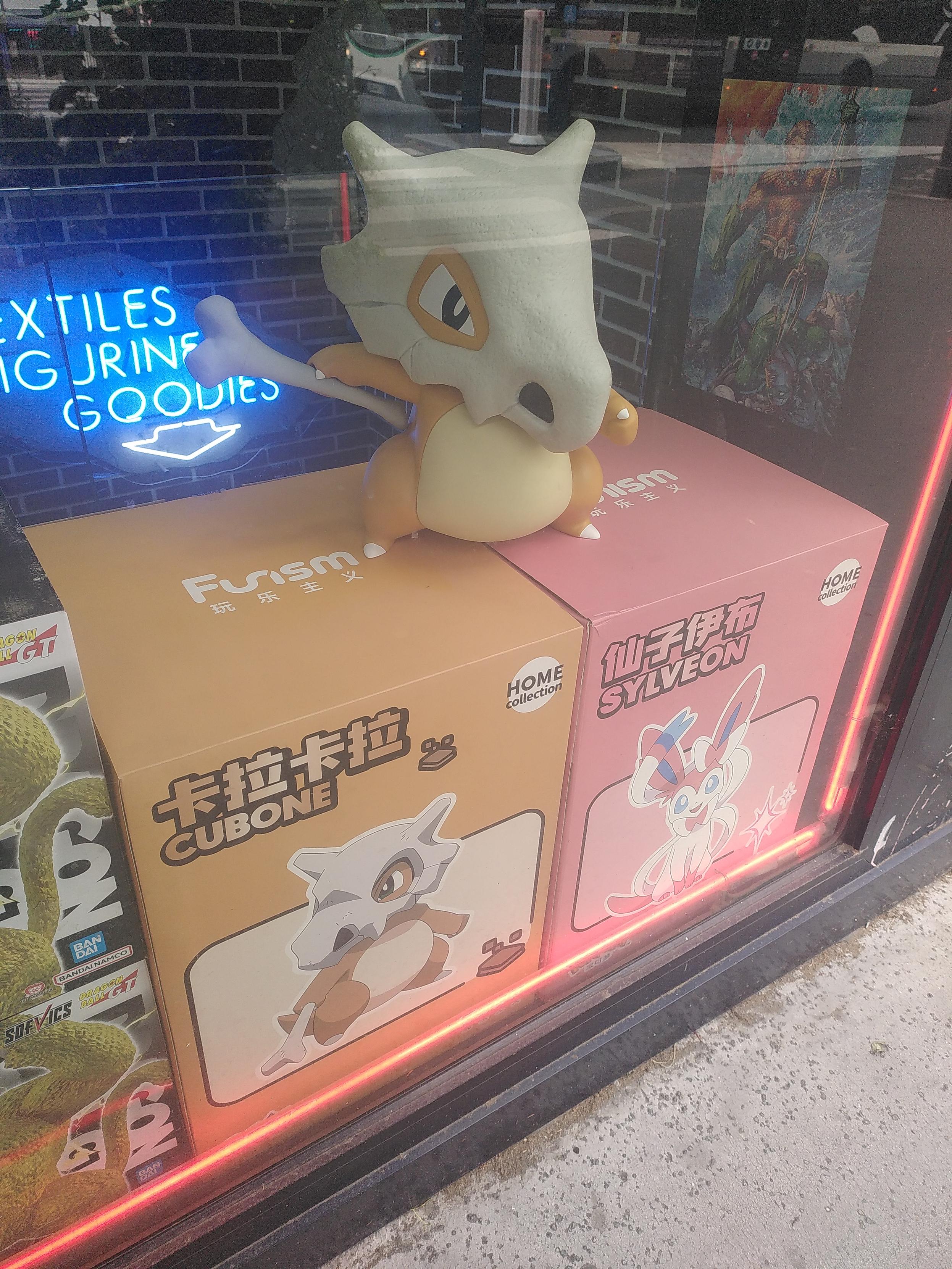 Photo of a shop's window. Inside is two boxes and a cubone standing on top of both. The thing is big, maybe bigger than life scale which is 40cm. Both boxes are the same cubic size and the second is pink with sylveon picture
