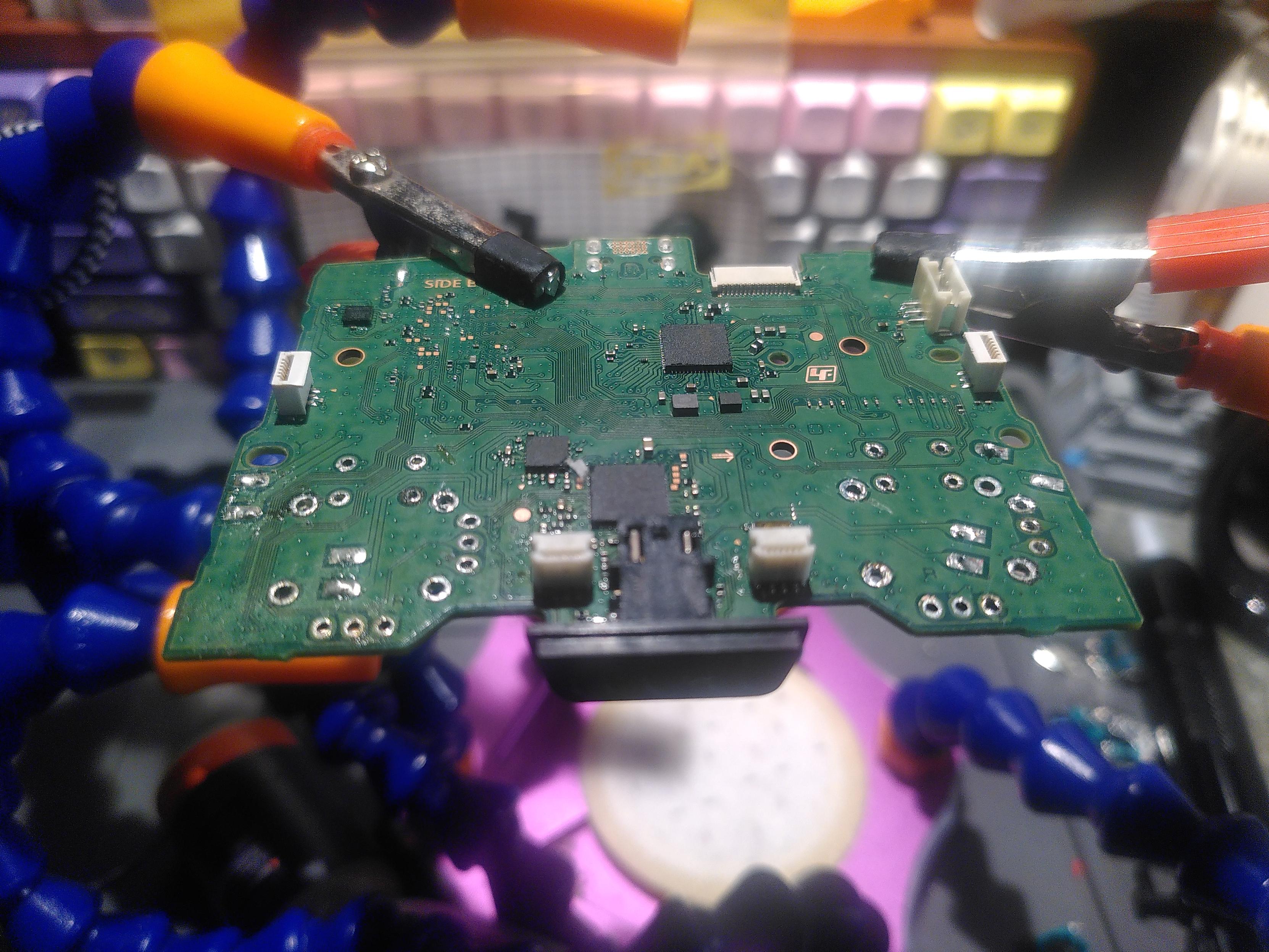 Photo of a green pcb. It's hold aloft by two claw attached to blue tentacles. There are 28 hole with silver edge more or less empty where two used to be. In the background is a blurry mechanical keyboard in a wooden shell