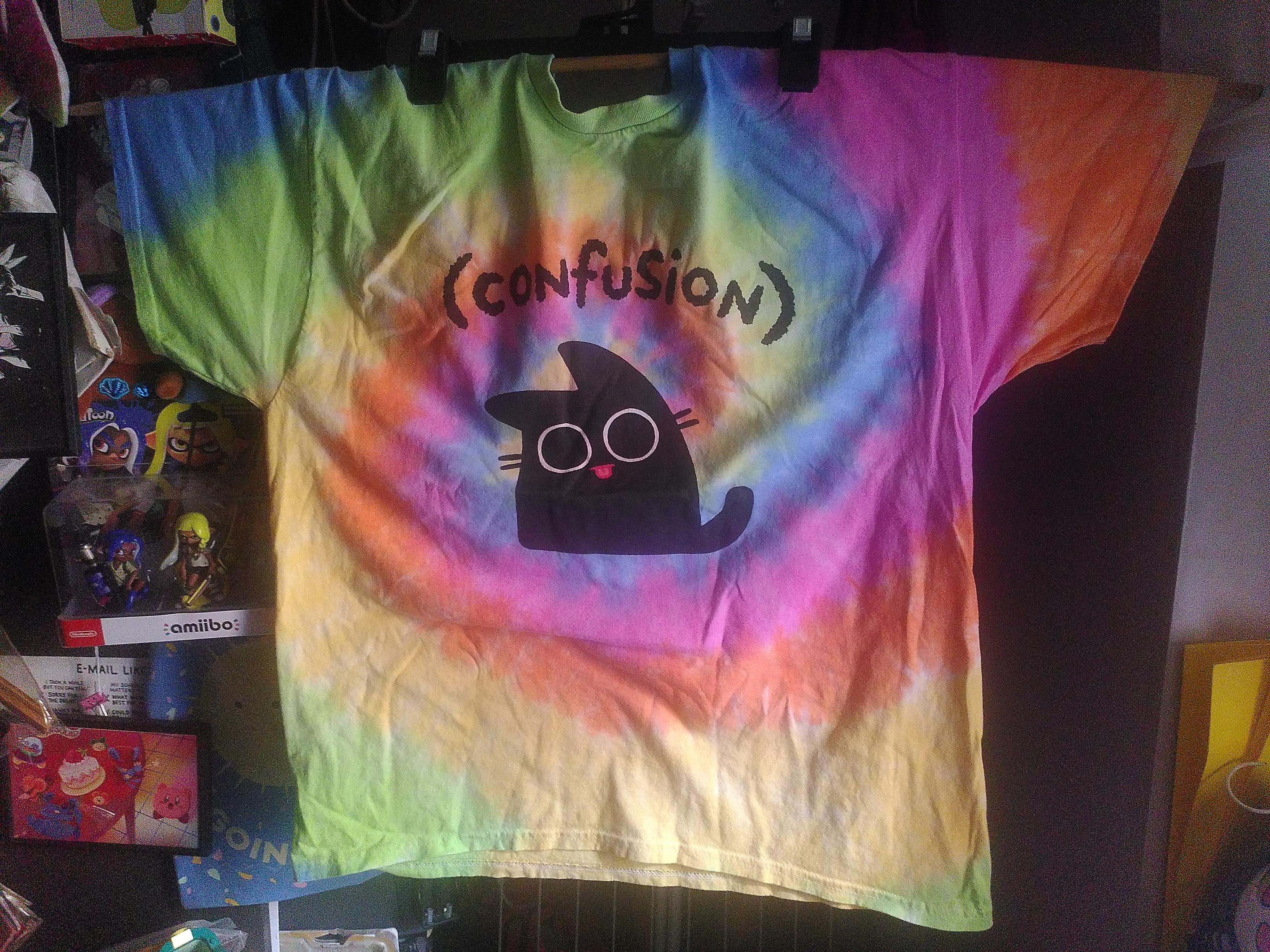 Photo of a tie-dye teesh. It's a colorful spiral, going blue, green, yellow, orange then pink. The color are warm ans soft. On the front, in the middle is a stylized black cat with giant eyes and a tiny red tongue going out. Above the cat is written in black (confusion). It's from Li Chen @exocomics@mastodon.world store