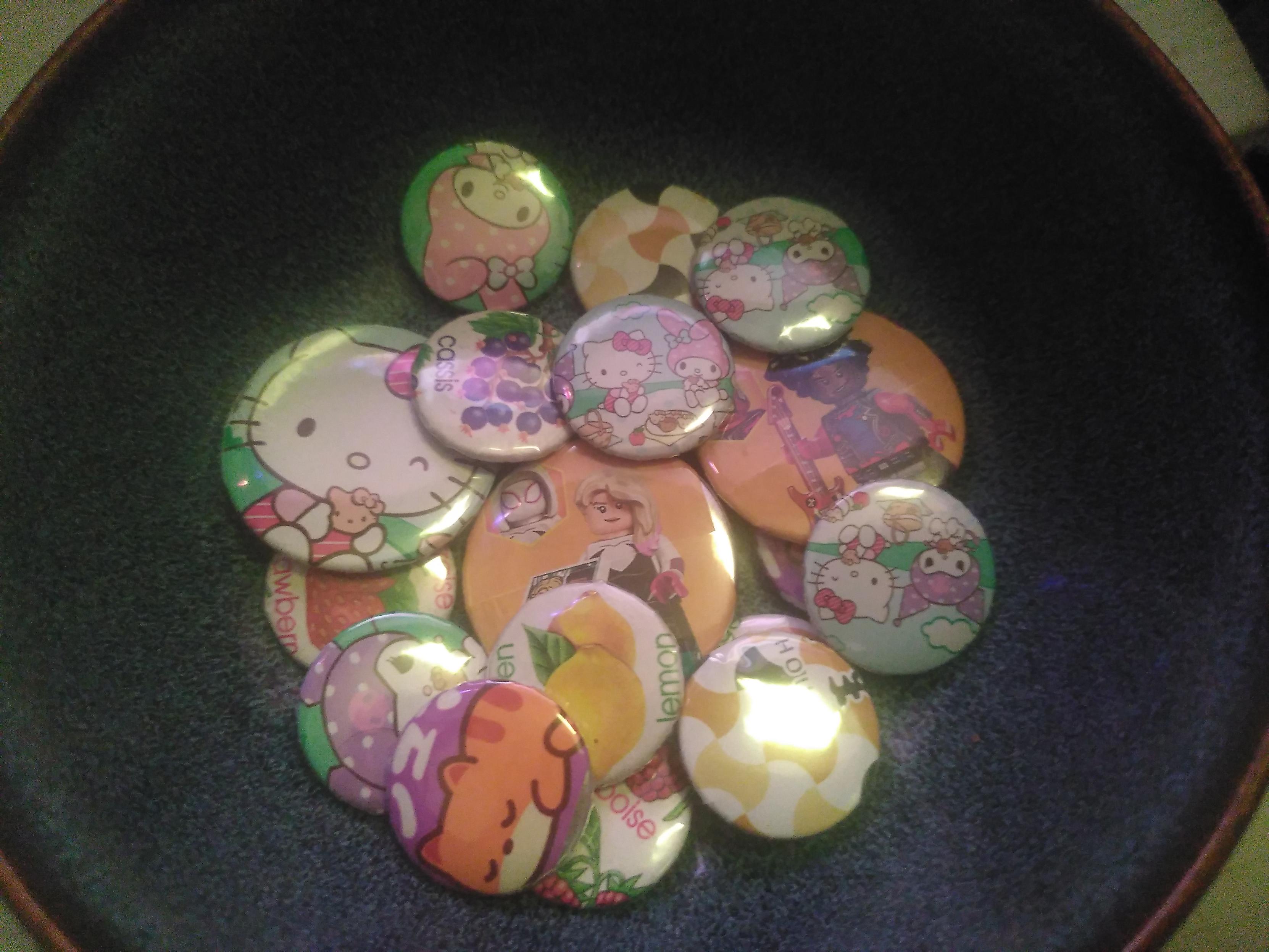 Photo of a bunch of button badge in an ikea bowl. The bowl is blue with lot of black speckles and a brown edge. Two of the badges have lego spider minigurines, (spider gwen and punk spiderman) (design salvaged from lego minifigurine blind box); a bunch have sanrio character like hello kitty (design salvaged from cookies); 5 have drawing of fruit with their name (blackcurrent, orange, lemon, strawberry, raspberry) (design salvaged from candy wrapper); two have abstract geometric pattern in white, light brown and black (design salvaged from cookie wrapper maybe); two have sleeping orange kitty (salvaged from candy wrapper)