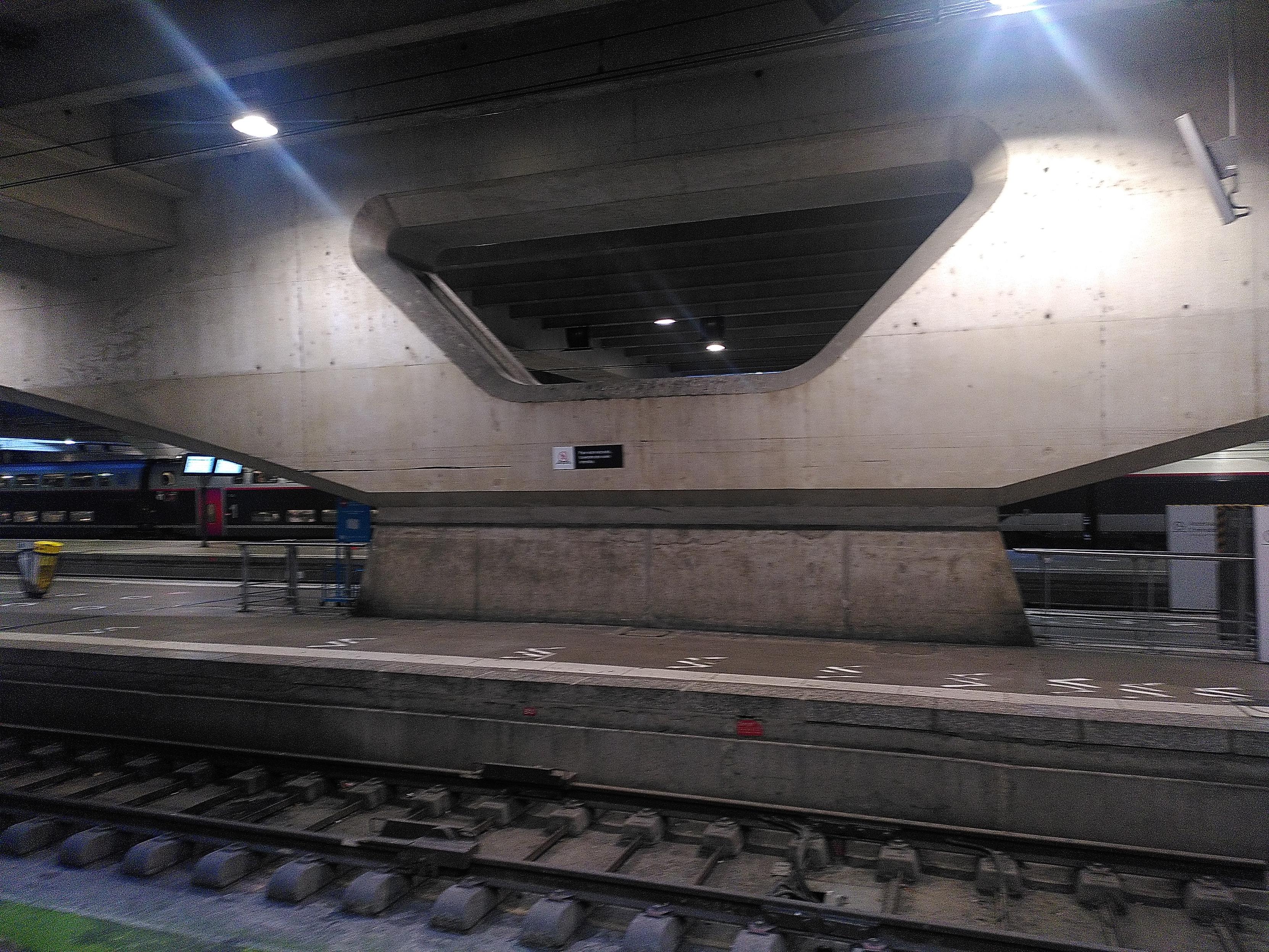 Photo of a concrete pillar inside montparnasse train station. It have a large base while the top is a V shape, the opening nearly as wide as the base and each branch ending also nearly as wide. At the bottom is a section of track made of metal rail and concrete support