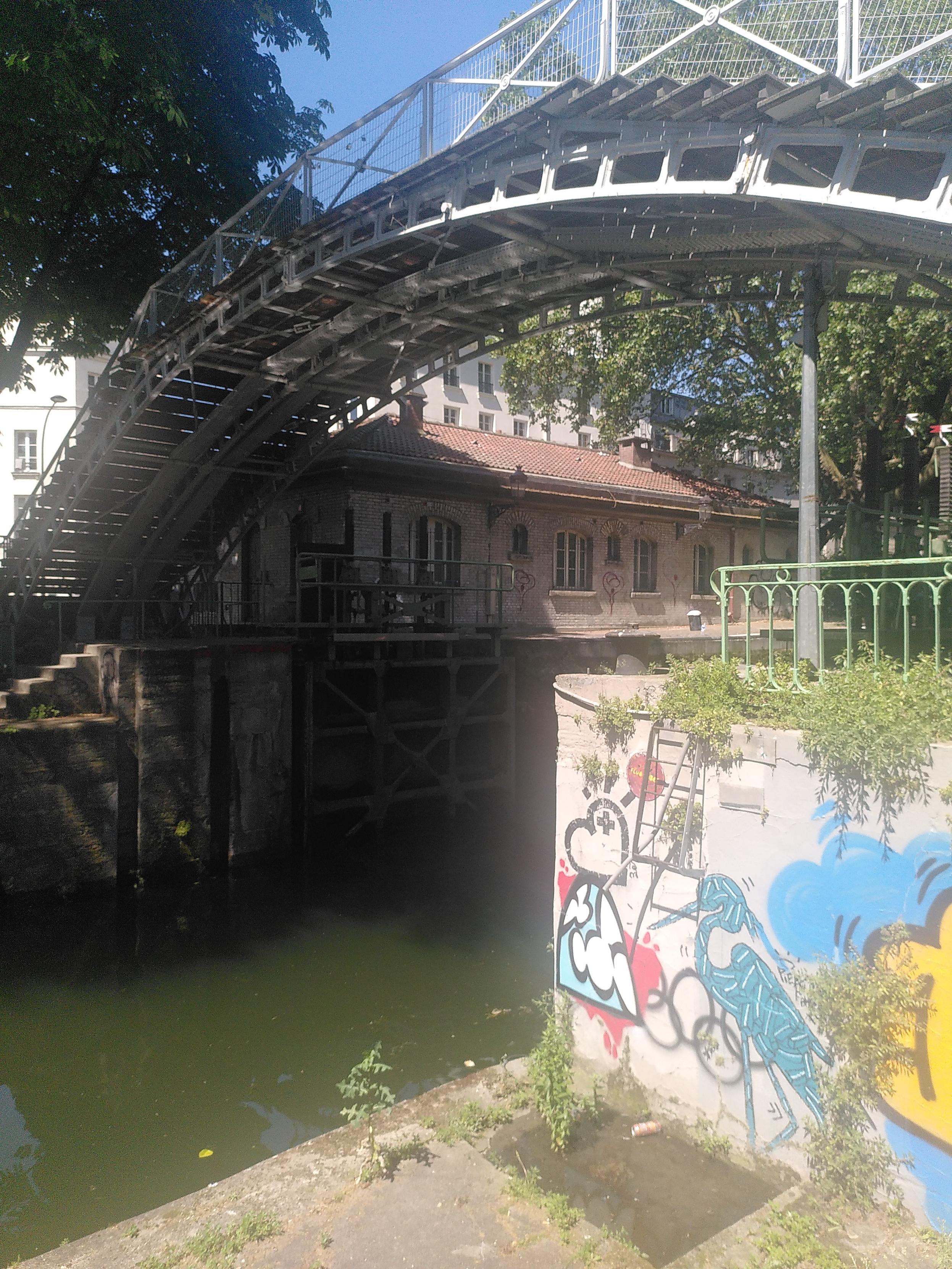 Photo of a bridge going over canal saint martin in paris. The sun is bouncing of the water and touching the bridge underside in moving pattern. The bridge is made of metal and wood, metal is painted light gray. The water is spinach smoothie. There are a few trees providing lot of shade and in the foreground is a pillar for the bridge made of concrete, covered in street art, with plenty sunlight