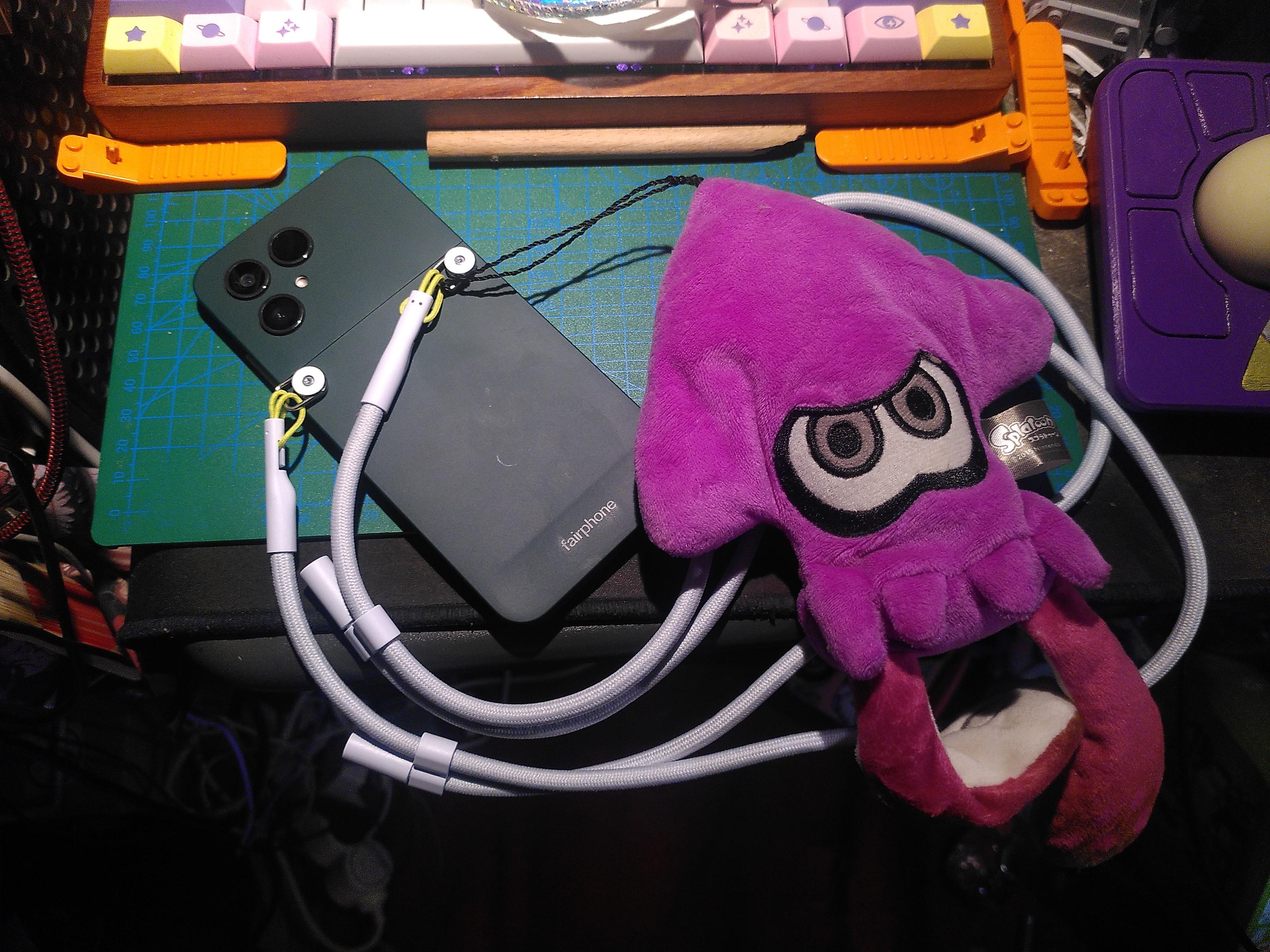 Photo of a forest green fairphone 6 with the lanyard accessory in cloud white and a splatoon plushie attached to the lanyard attachment point. The plushie is purple with white/gray eyes and white tentacle underside. It looks like a squid made if a arrow-like main body, as high as the phone, as width as the phone under the triangle of the arrow, plus two tentacle attached under that are 80% of the height if the phone