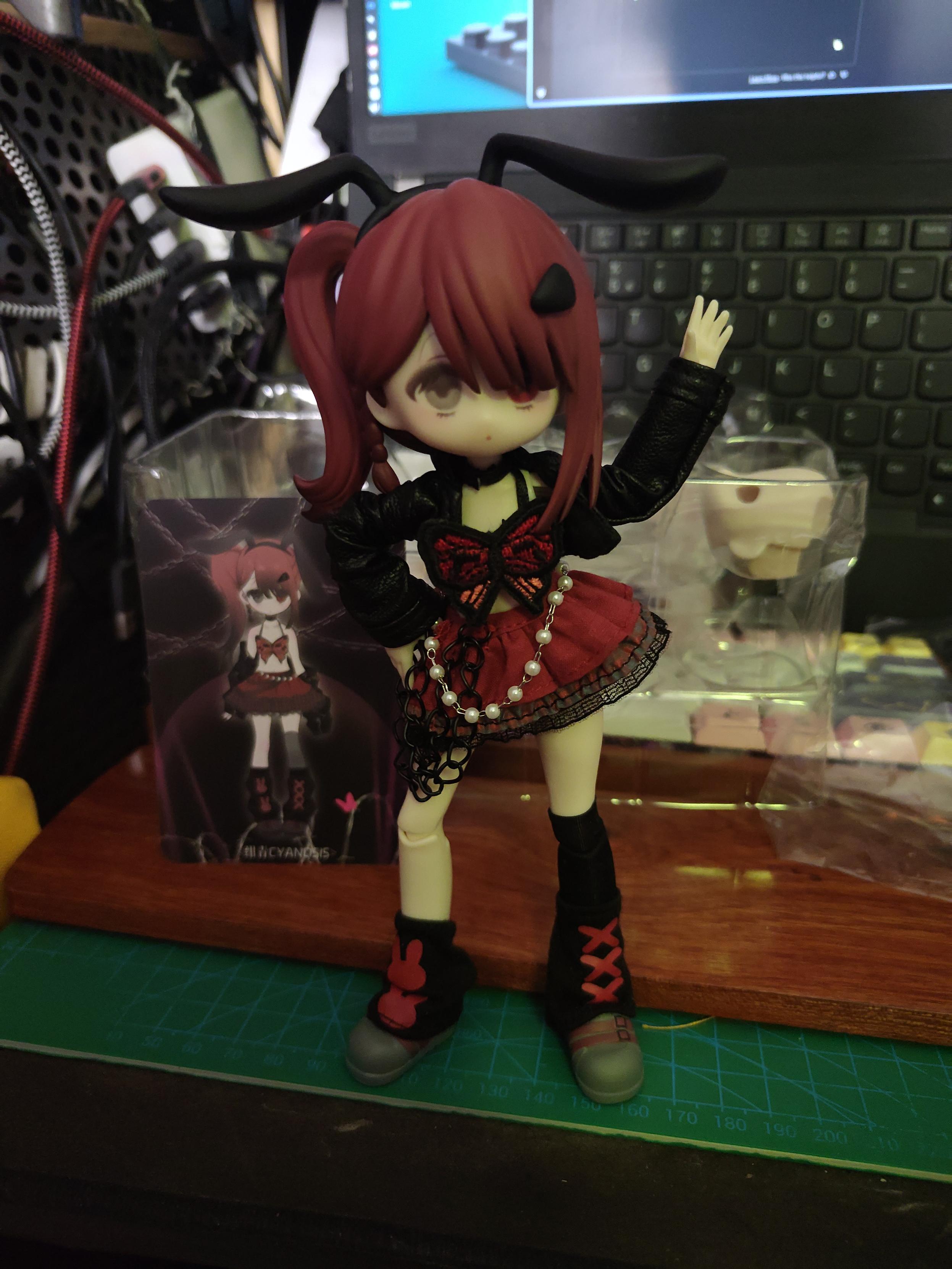 Photo of a ball joint doll standing on a desk. She has fair skin, plastic red hair in a complexe hairdo involving a side ponytail going down to her shoulder, a bang and a more stuff, black bunny ears, a black leather jacket, a crop top looking like a red butterfly with black outline, a rust red mini skirt with black dentelle, black chain and silver and pearl chain, one knee length black sock, two black leg warmer, one with two red bunny head motif and the other with three red x motif, gray plastic shoes in a shape that remind of leather shoes, with painted brown strap. She has one hand on her hips and is waving the other. 