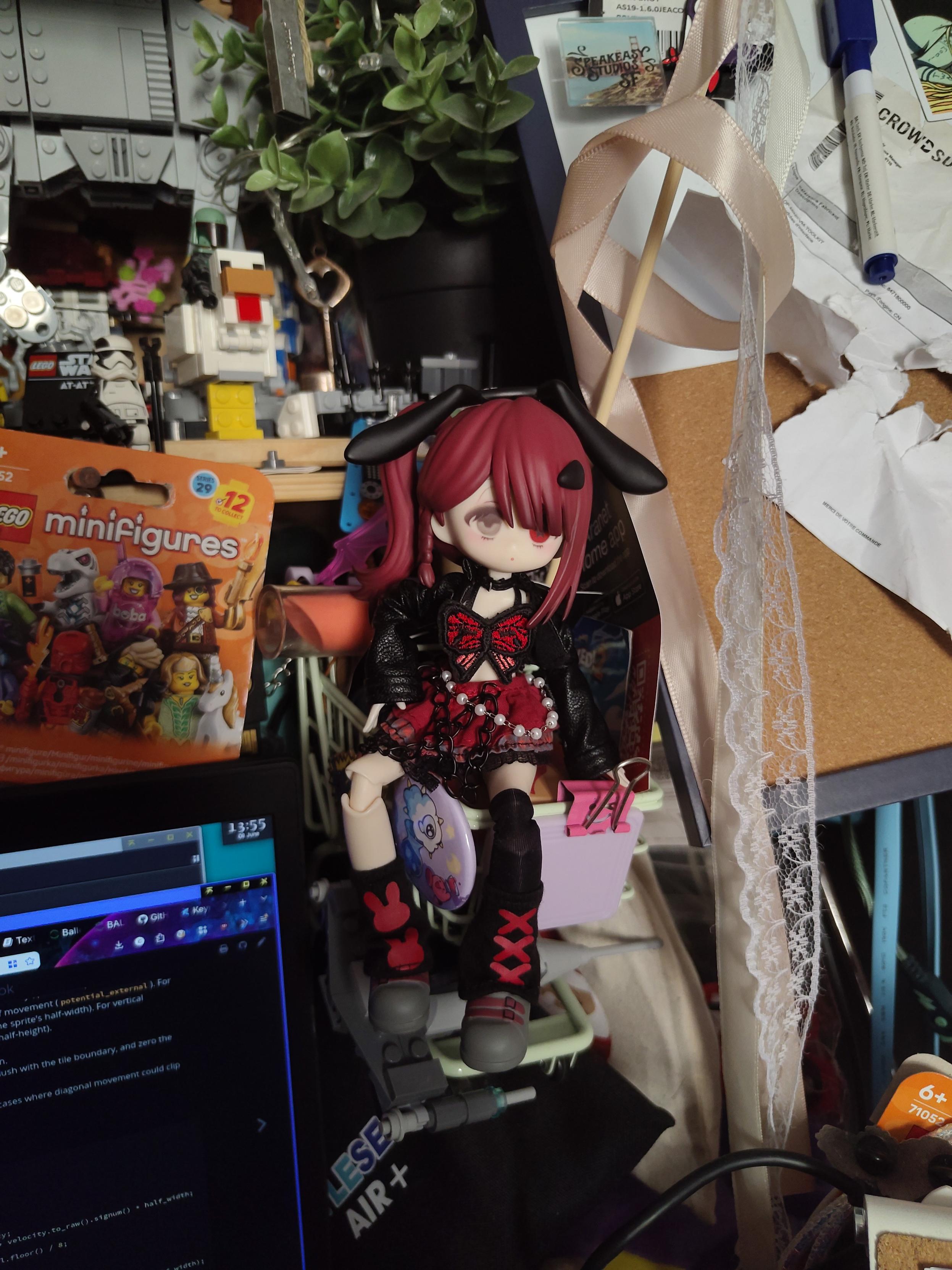 Photo of a ball joint doll wearing clothings, sitting on a small shopping cart, suspended above my desk