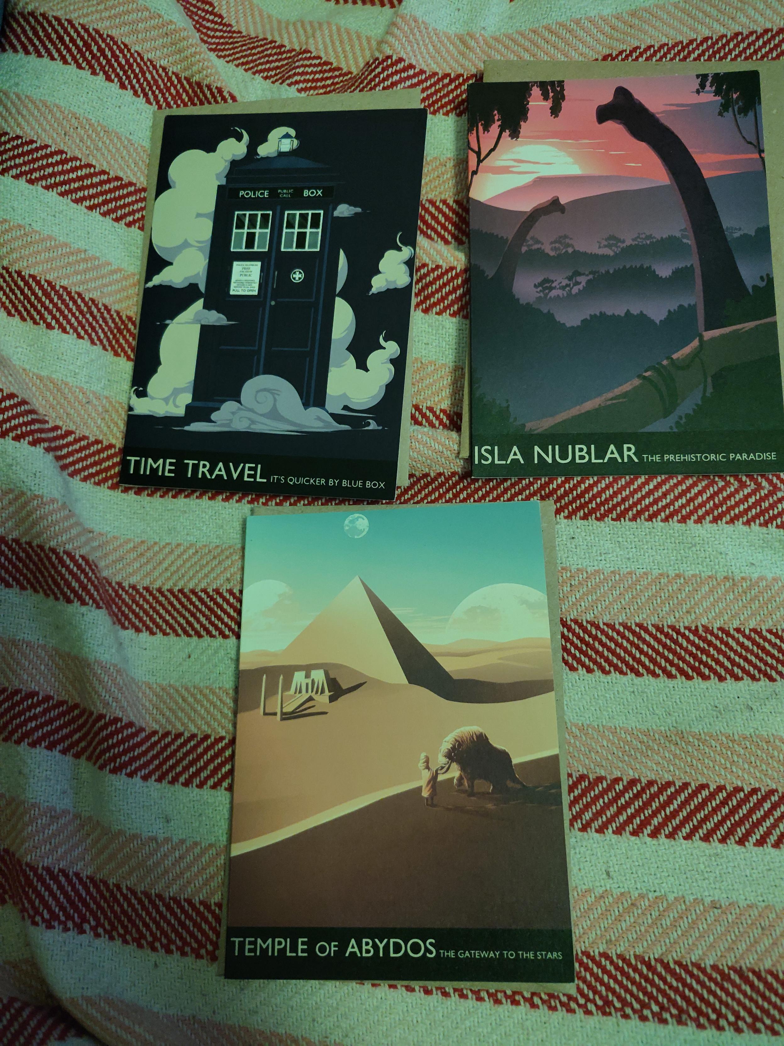 Photo of 3 postcard on some clothing. Each postcard have an illustration for a piece of pop-culture, namely doctor who, jurassic park and stargate, with under it a title and a catch phrase. the first one show a dark blue phonebooth in some white smoke/most, with 