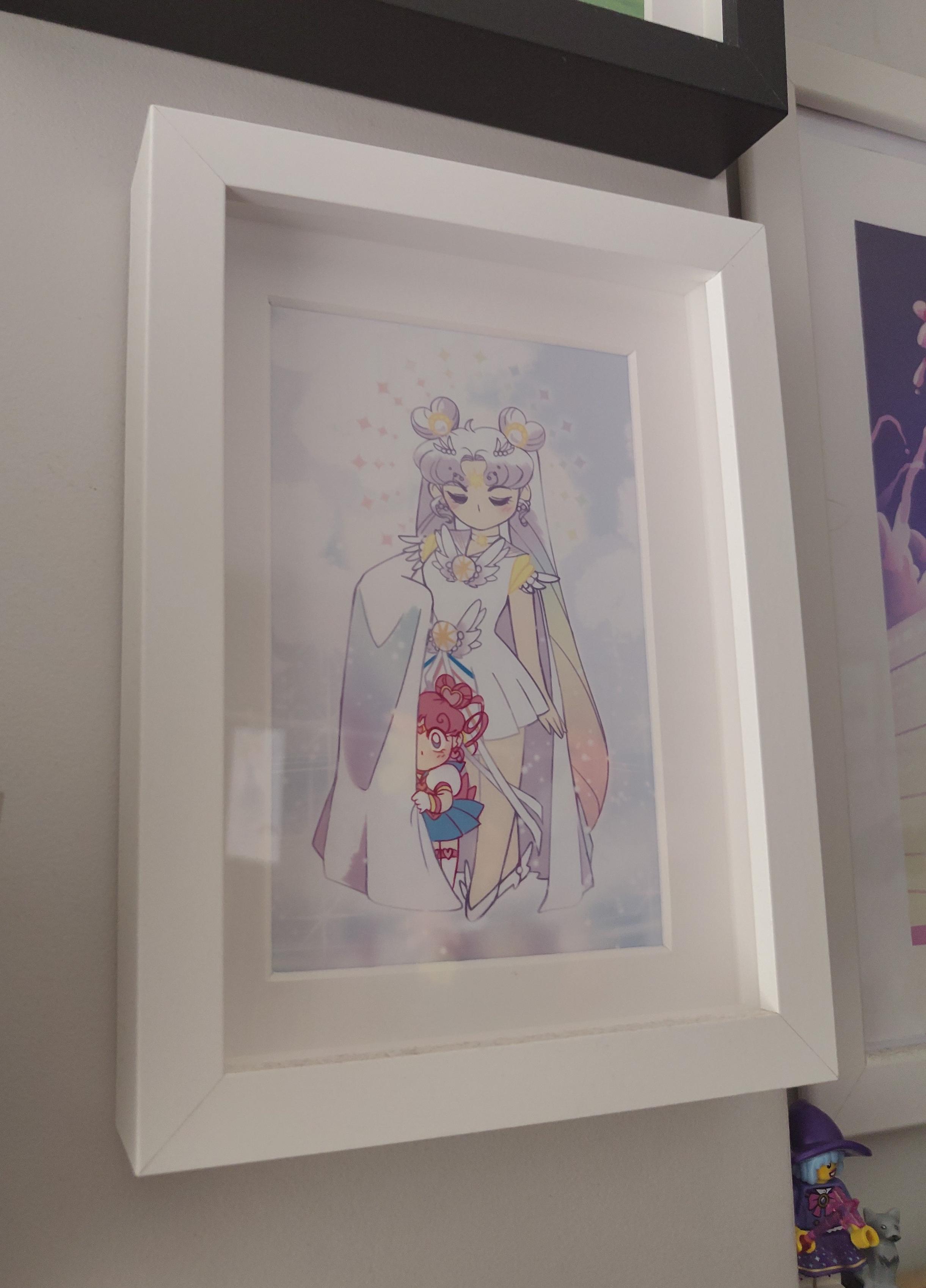 Photo of a framed mini-print from Doodlemancy. The white frame has a bit of depth, with the plastic maybe a centimeter from the paper, with a white border around the print. The illustration is mostly white with pastel shade of blue, purple and peach, featuring a magical girl  drapping her right arm with her cape. Half hiding behind that part of the cape is a chibi sailor moon, barely the height of the protective figure's hip, with royal blue and crimson pink hair.