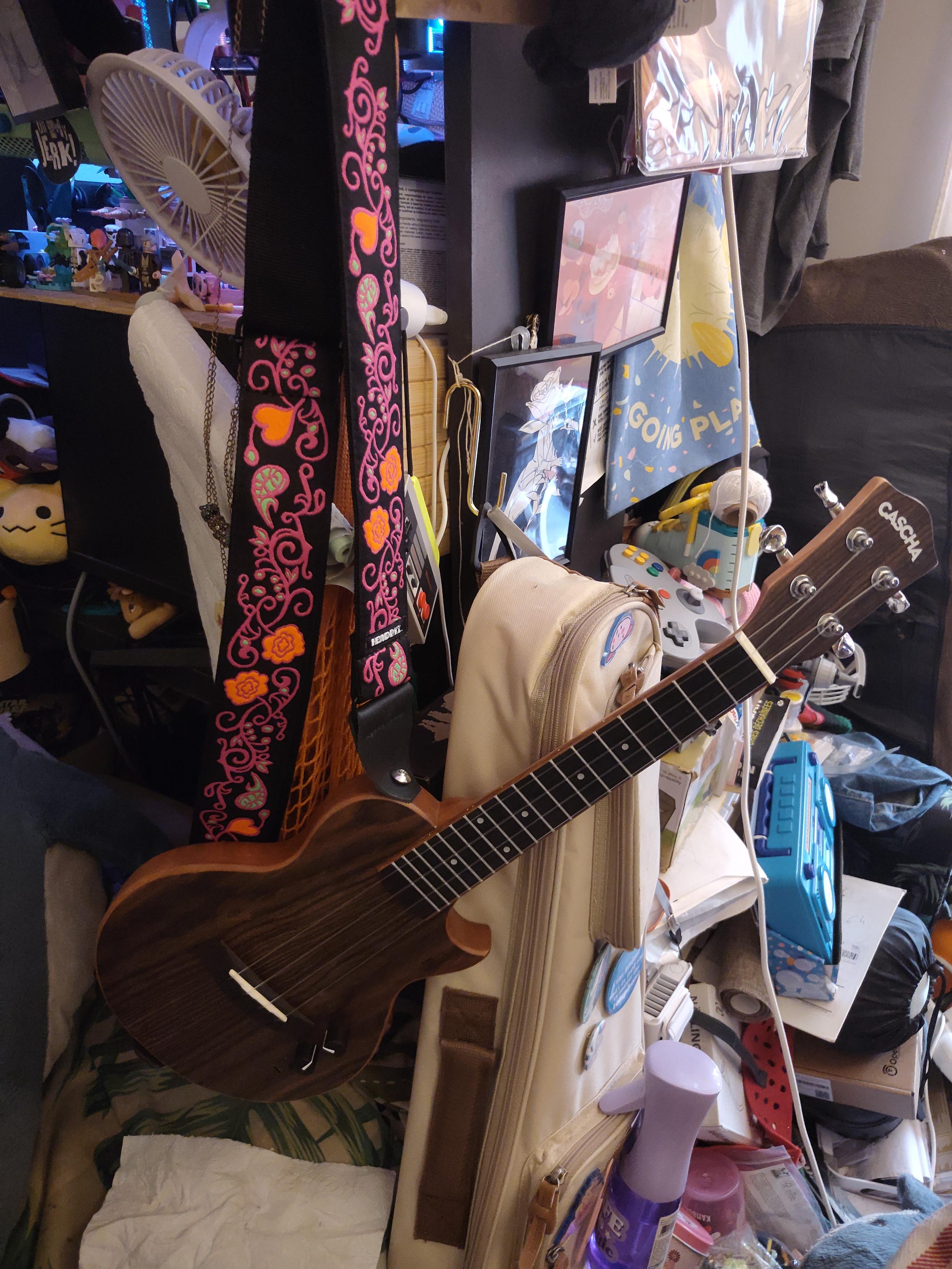 Photo d'un ukulele electrique avec une bretelle de guitare. Le ukulele est en bois assez sombre et la bretelle noire avec des motifs genre lianes et feuilles roses parfois remplie en vert et des fleur rempli orange. Le ukulele est suspendu par la bretelle et il y a beaucoup de bazar derriere.