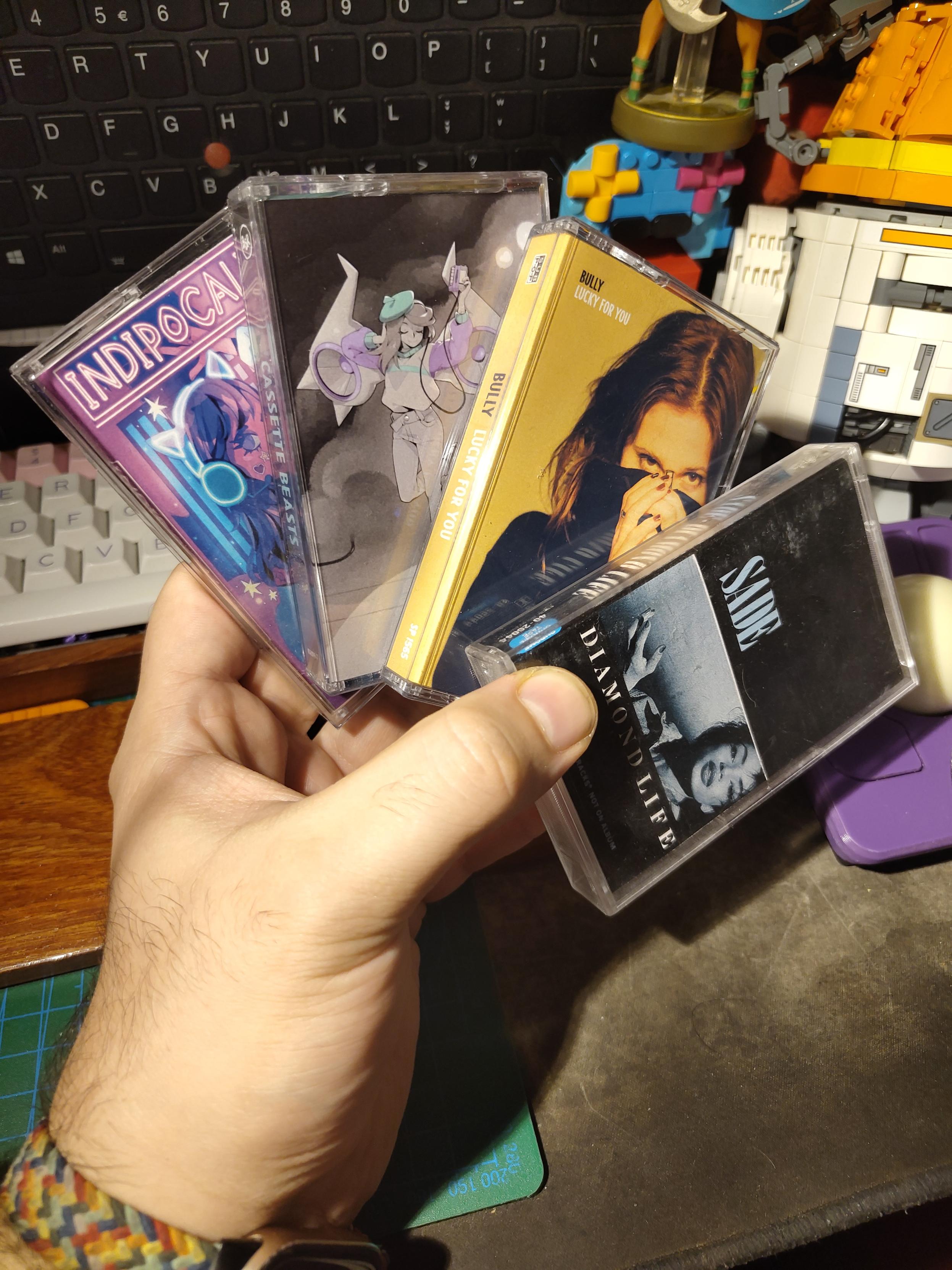 Photo of my hand holding 4 cassette tape box, fanned out so illustrations are visible; 
first one is Diamond Life by Sade, with a mostly black illustration cut by a black and white portrait of sade take up a third of the space, below the center; Second one is Lucky for You by Bully, the illustration is a photo of a white woman with brown hair holding a black hoodie collar up to her eyes, hiding half of her face. The hoodie cover half of the illustration, the background is a soft yellow; Third box is Cassette Beast background music. The illustration is a black and white drawing of a young woman listening to a tape player. She has wings with large rotor and a couple of large spikes. There are pastel purple accent on the rotor center, rim, the tape player and the woman sweatshirt arms. There are pastel green accent on the woman berret, sweatshirt arms' rim, a strip in the center of the sweatshirt and the sweatshirt collar; The fourth box is indipocalypse 016 and contain not a tape but a usb flashdrive with a compilation of indie game. The illustration is a digital drawing in pink and blue, featurinf a young woman with flower tatoo, a short skirt, a top showing plenty cleavage, large sleeve on her arms and a headset + mic with cat ears and neon outline. She is sorrounded by star and skyscrapper are visible in the back.
