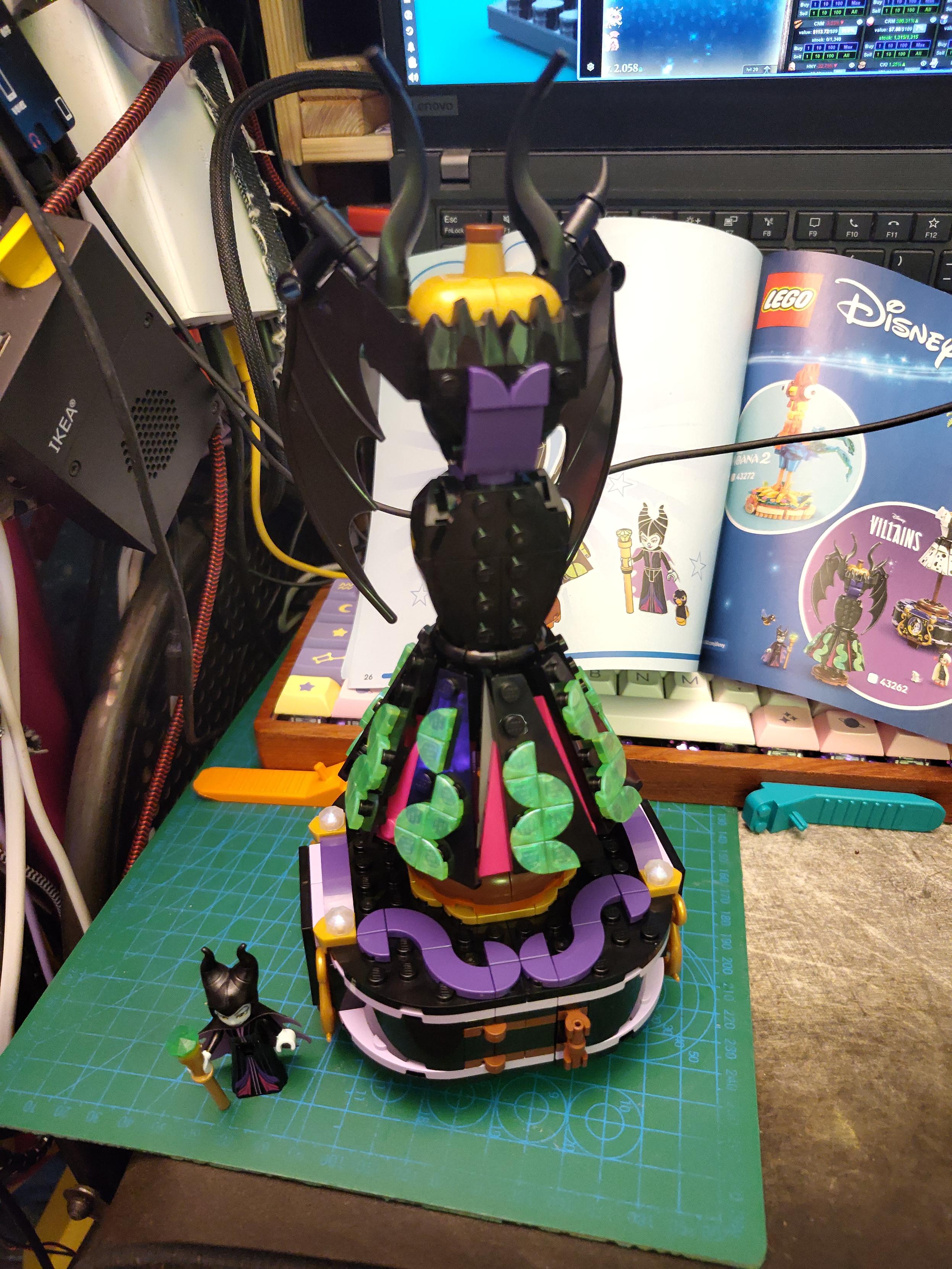 Photo of some lego. It's Maleficient dress on a wooden mannequin, next to smoll maleficient minidoll. The dress have wasp is mostly black with a purple piece on the front middle from waist to underboob, red pleat and curvy shiny emerald  curvy pattern on the part below the knee. The robe itself has black, folded down wings, a wasp weight and a bottom part that get thinner around the knee before getting wider